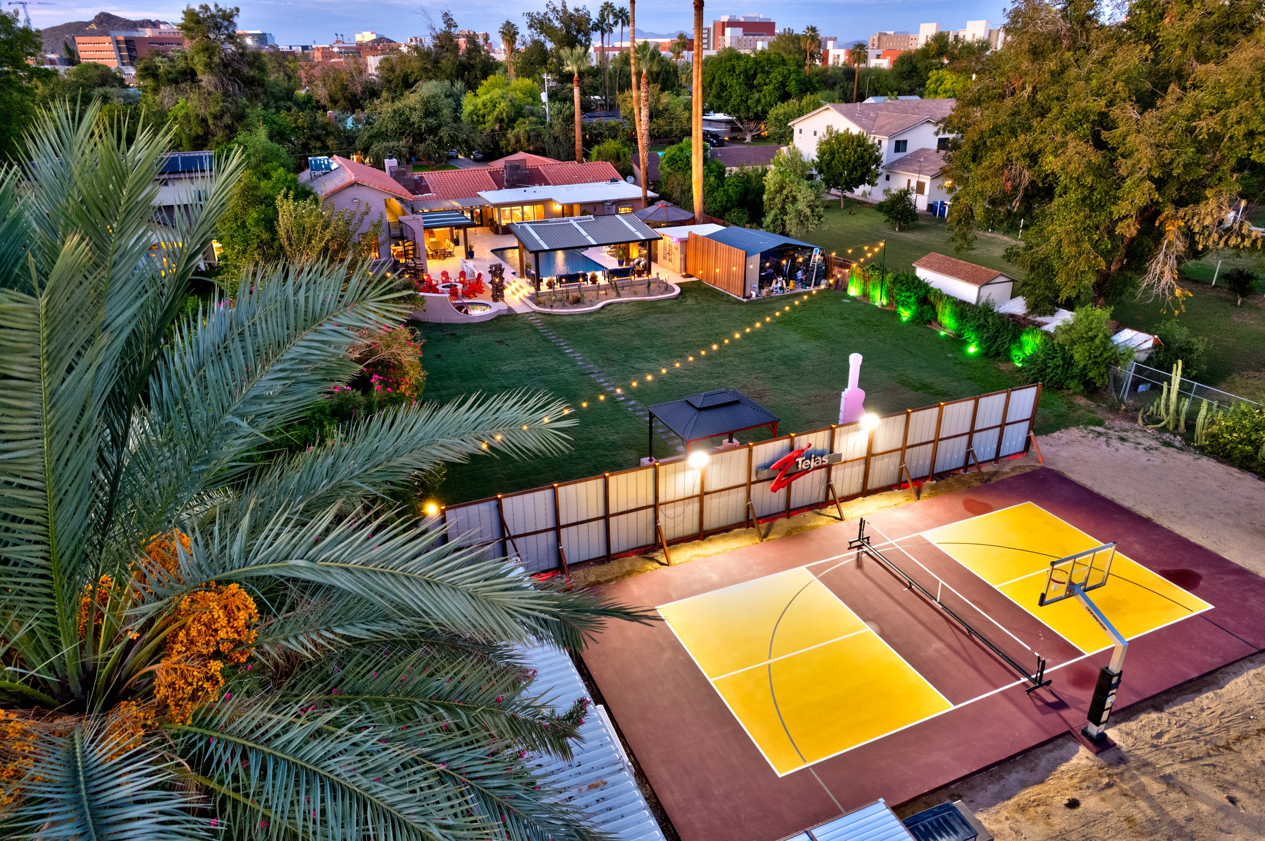 Twilight aerial view of a Tempe backyard with a swimming pool, ramada, and illuminated tennis and basketball court.