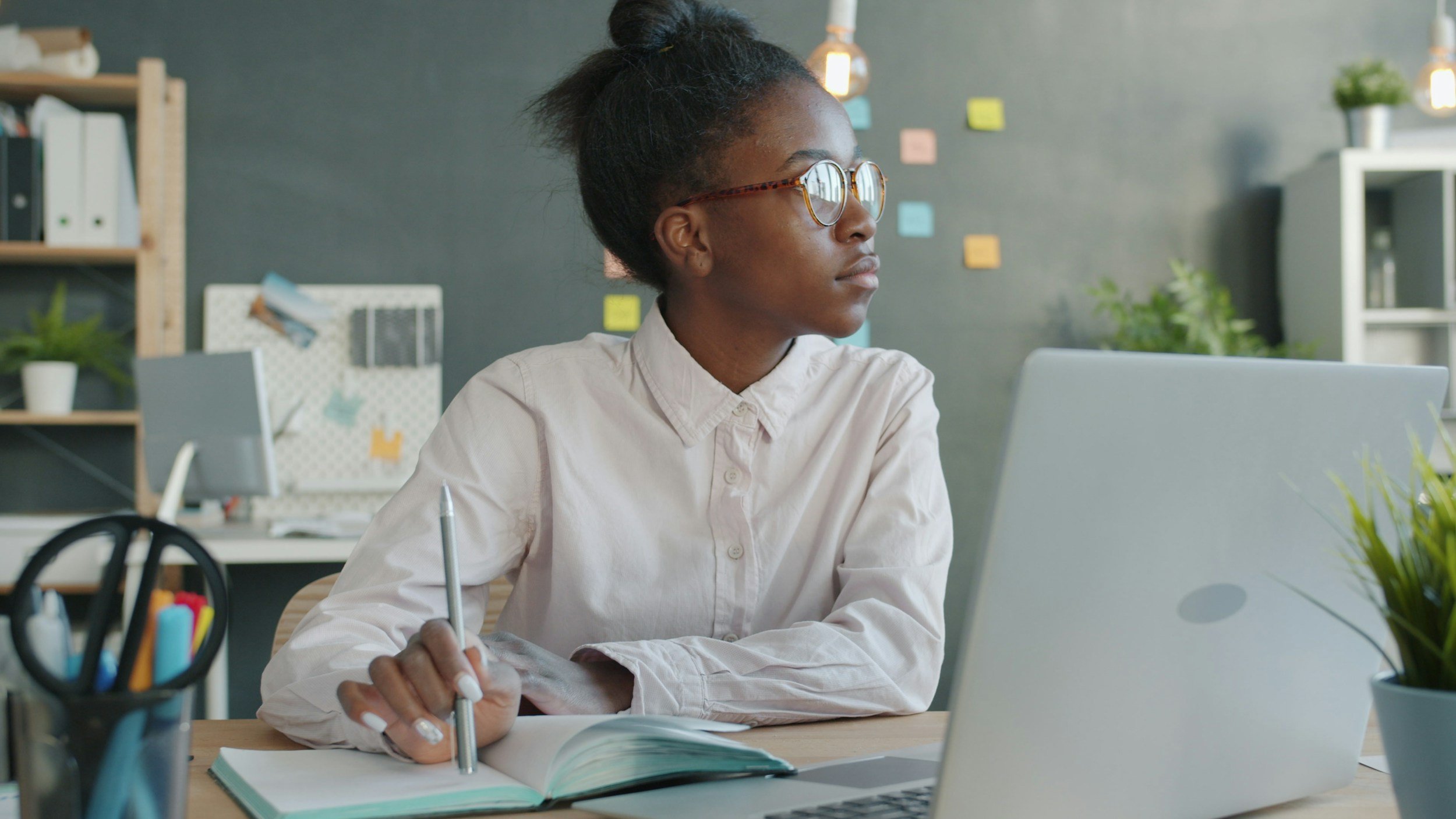 a young black woman working at a laptop in an office space, stares out the window
