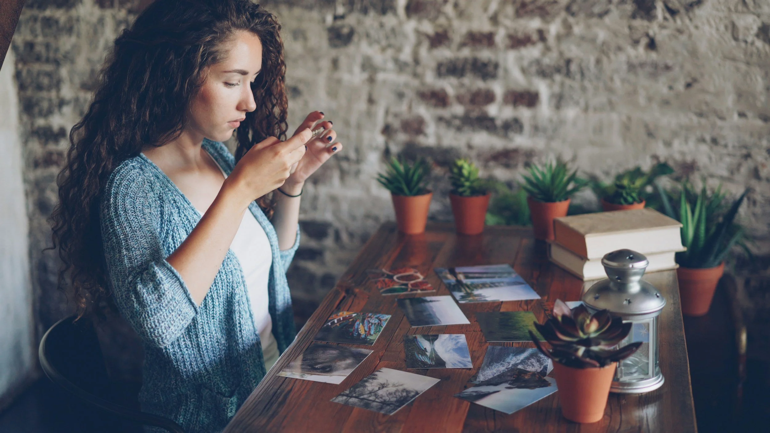 a caucasian woman in a blue cardigan stands in front of a table. there are photographs covering the table and she is holding a photograph up to her face and she is studying it.