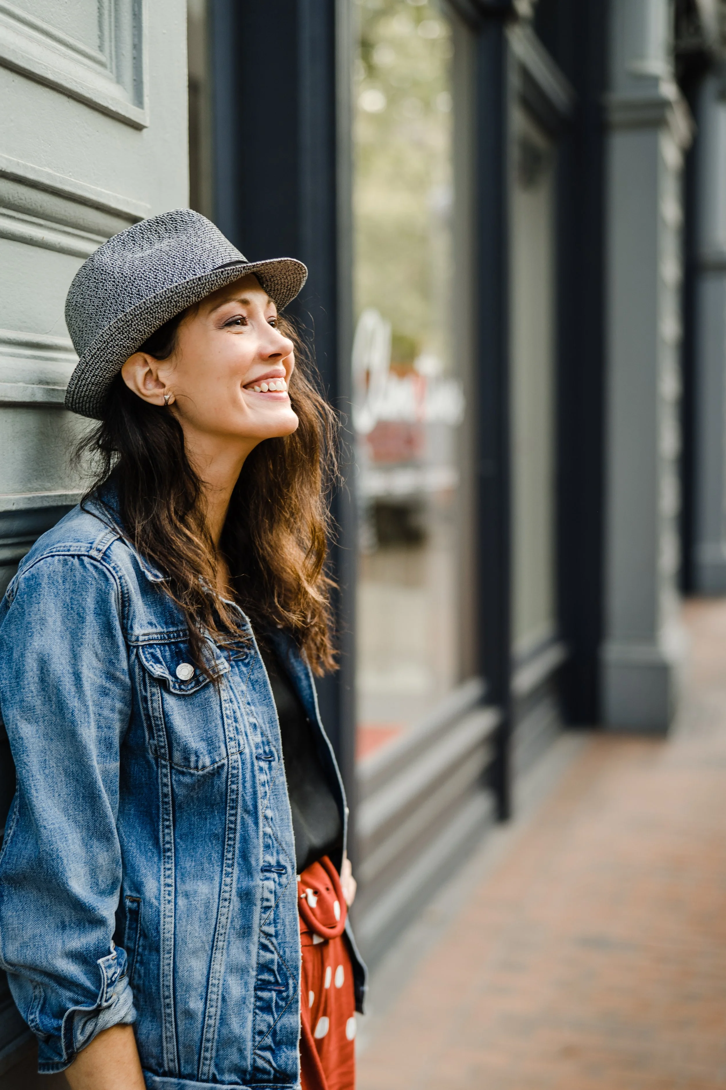 a portrait of the therapist, a woman in a fedora, jean jacket leaning against a wall in profile, smiling and looking out