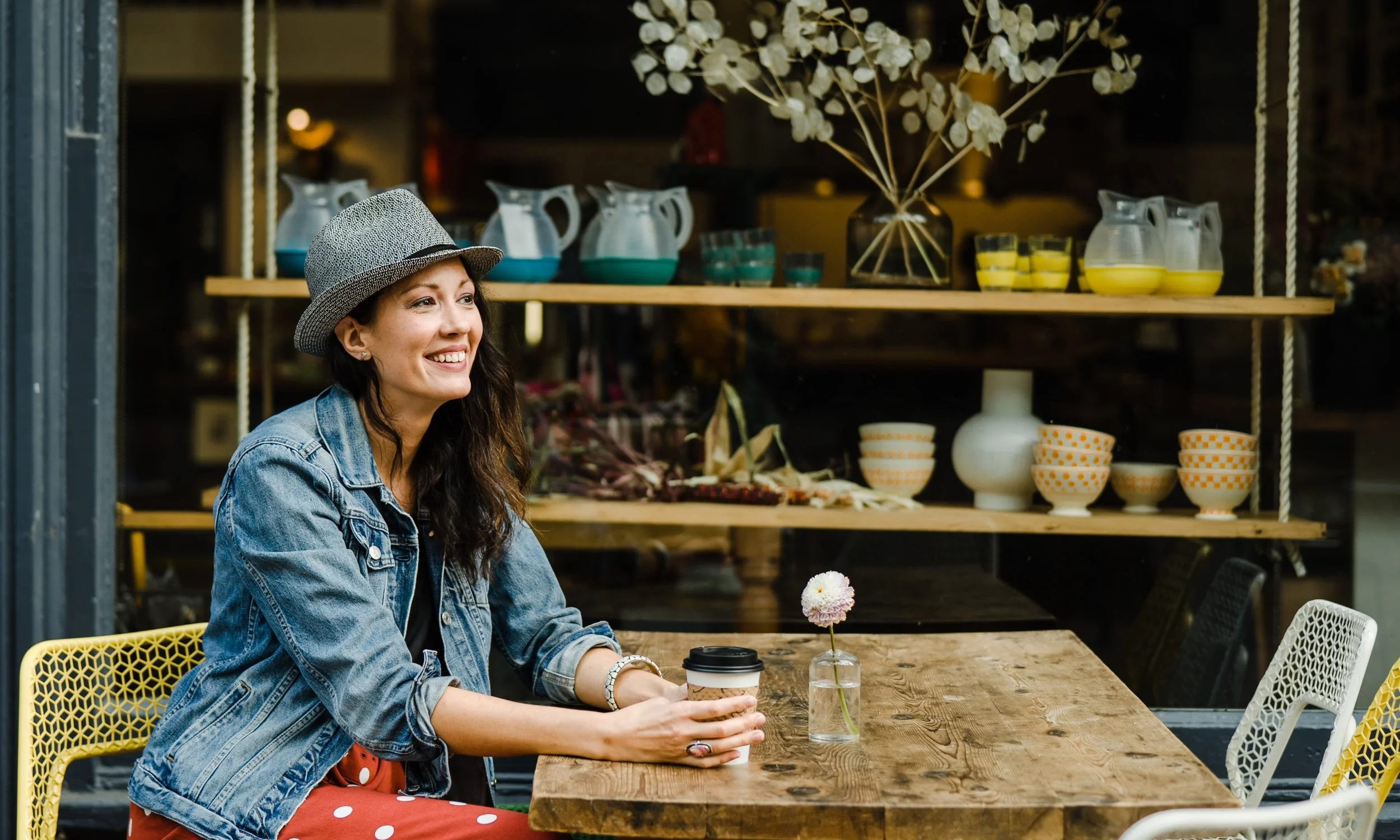 portrait of the therapist - a woman with wavy hair and a fedora - at a table with a coffee looking across the table while smiling