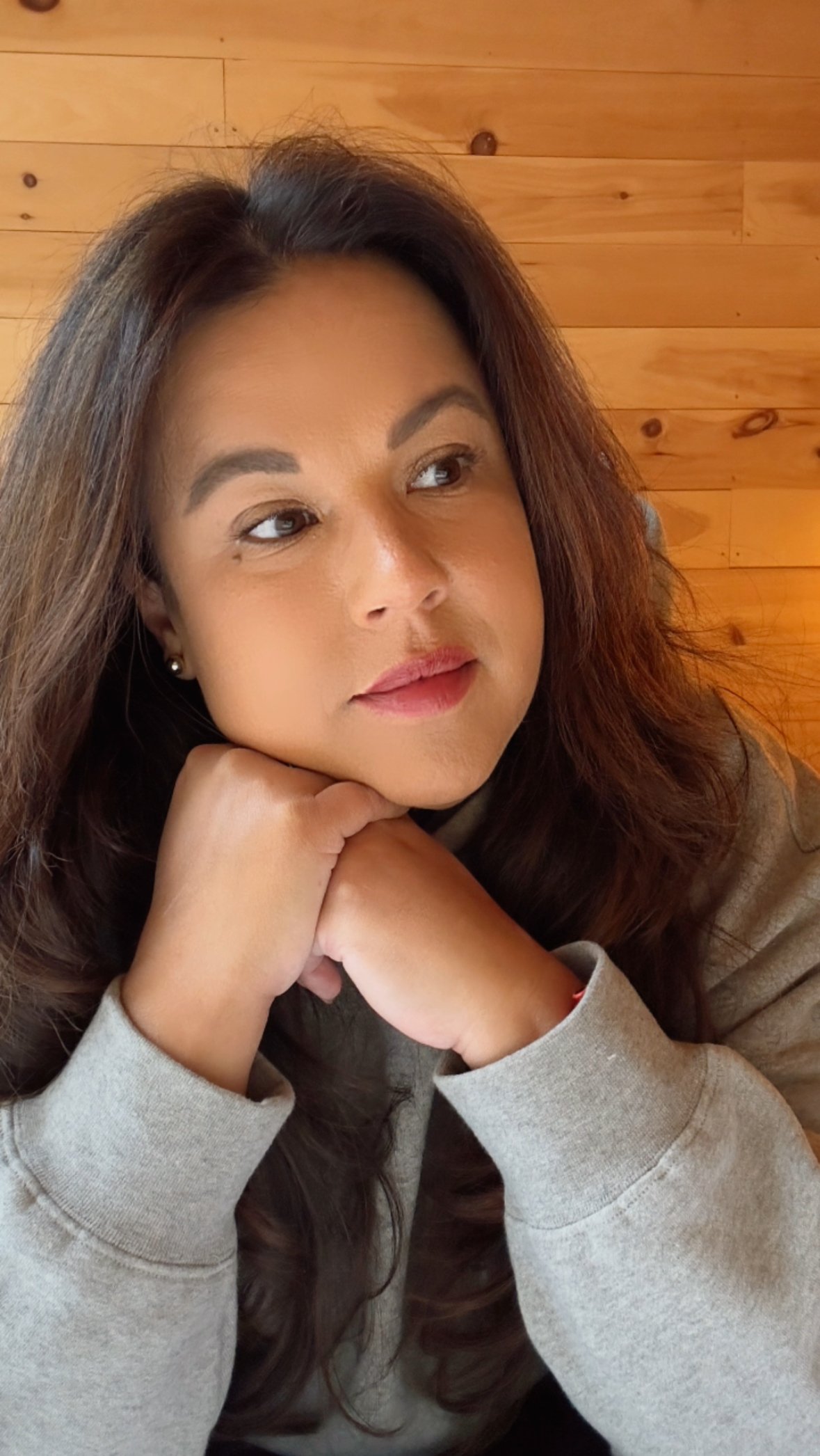 A woman with long brown hair resting her chin on folded hands, gazing thoughtfully to the side, against a wooden wall background.