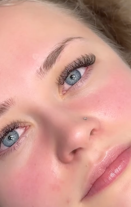 Close-up of a woman's face showing her blue eyes, natural eyebrows, long eyelashes, a small nose piercing, and lightly glossed lips.