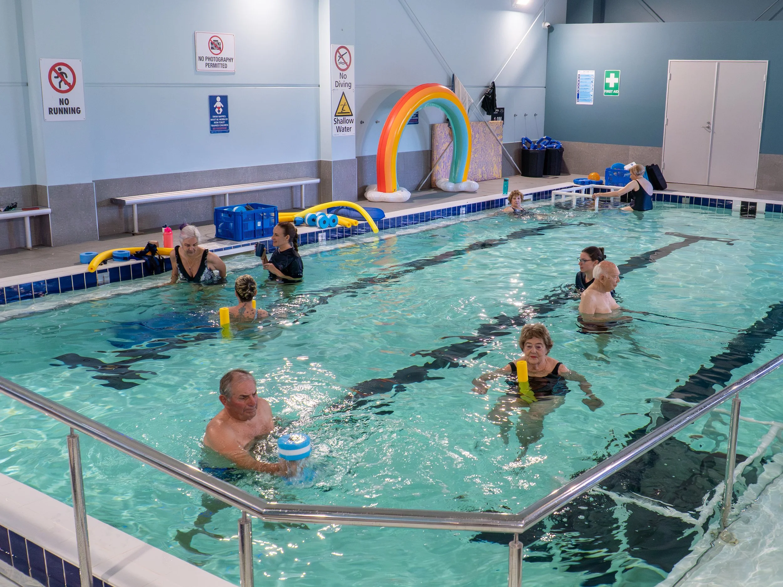 An indoor swimming pool with several elderly and young people practicing water exercises. Some are using pool noodles, and a staff member assists near the edge. There are various pool safety and no diving signs on the wall.
