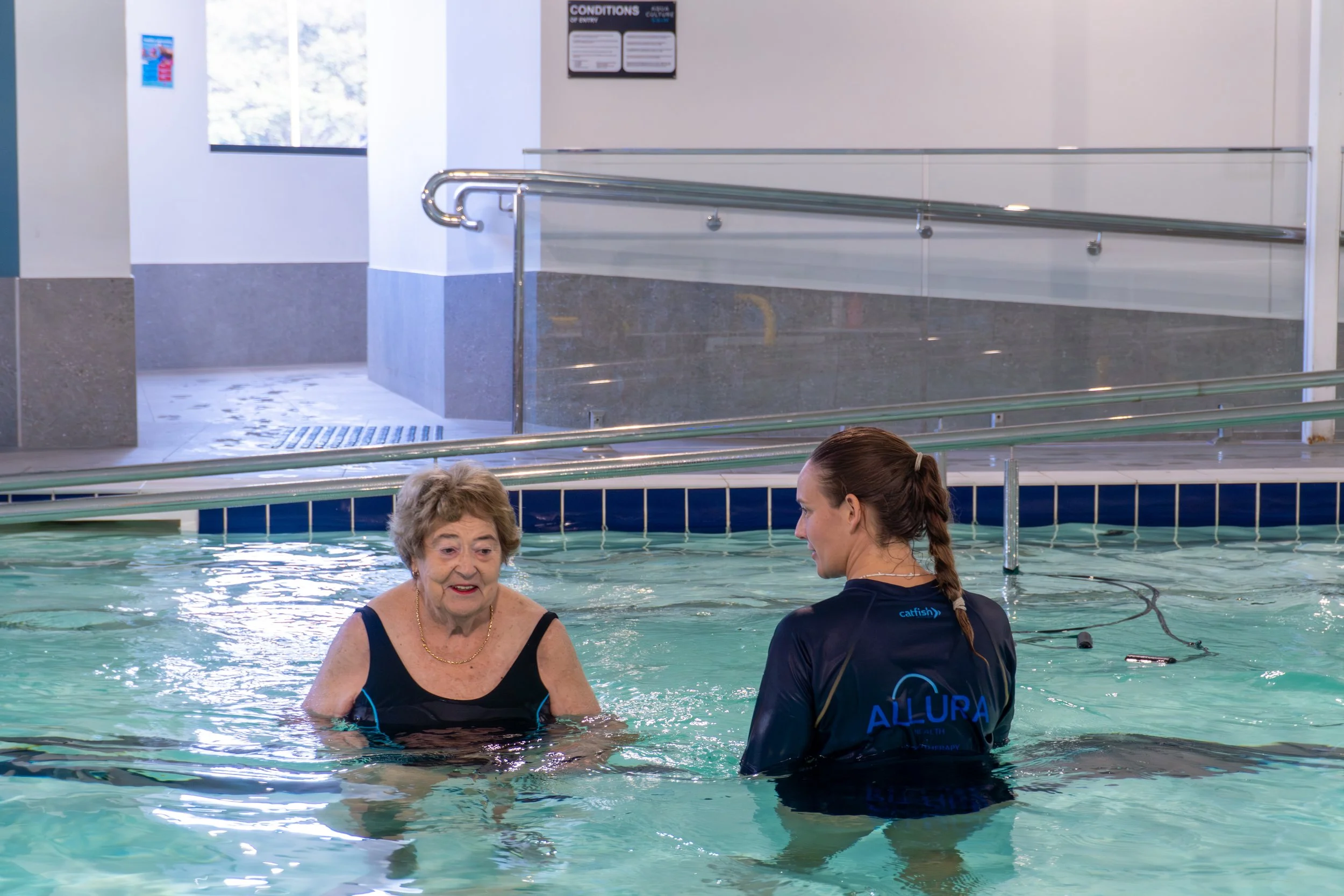 An elderly woman and a swim instructor having a conversation in a therapy pool at a rehabilitation center.