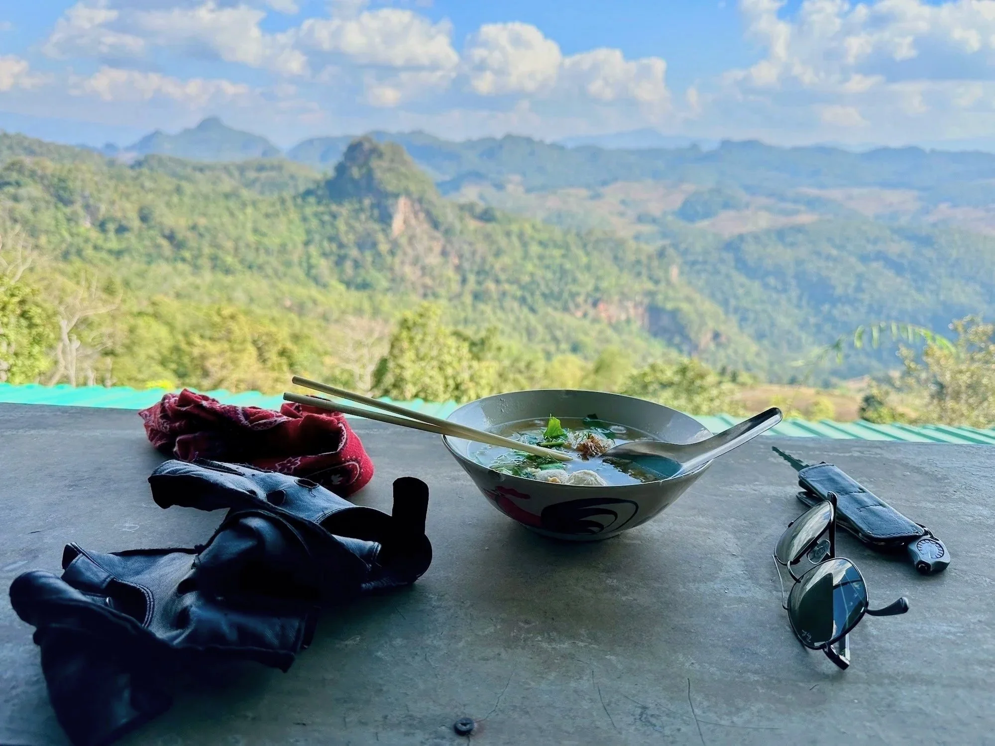 A bowl of noodles along with the author’s biking gloves and sunglasses on a table with a stunning mountain view behind.