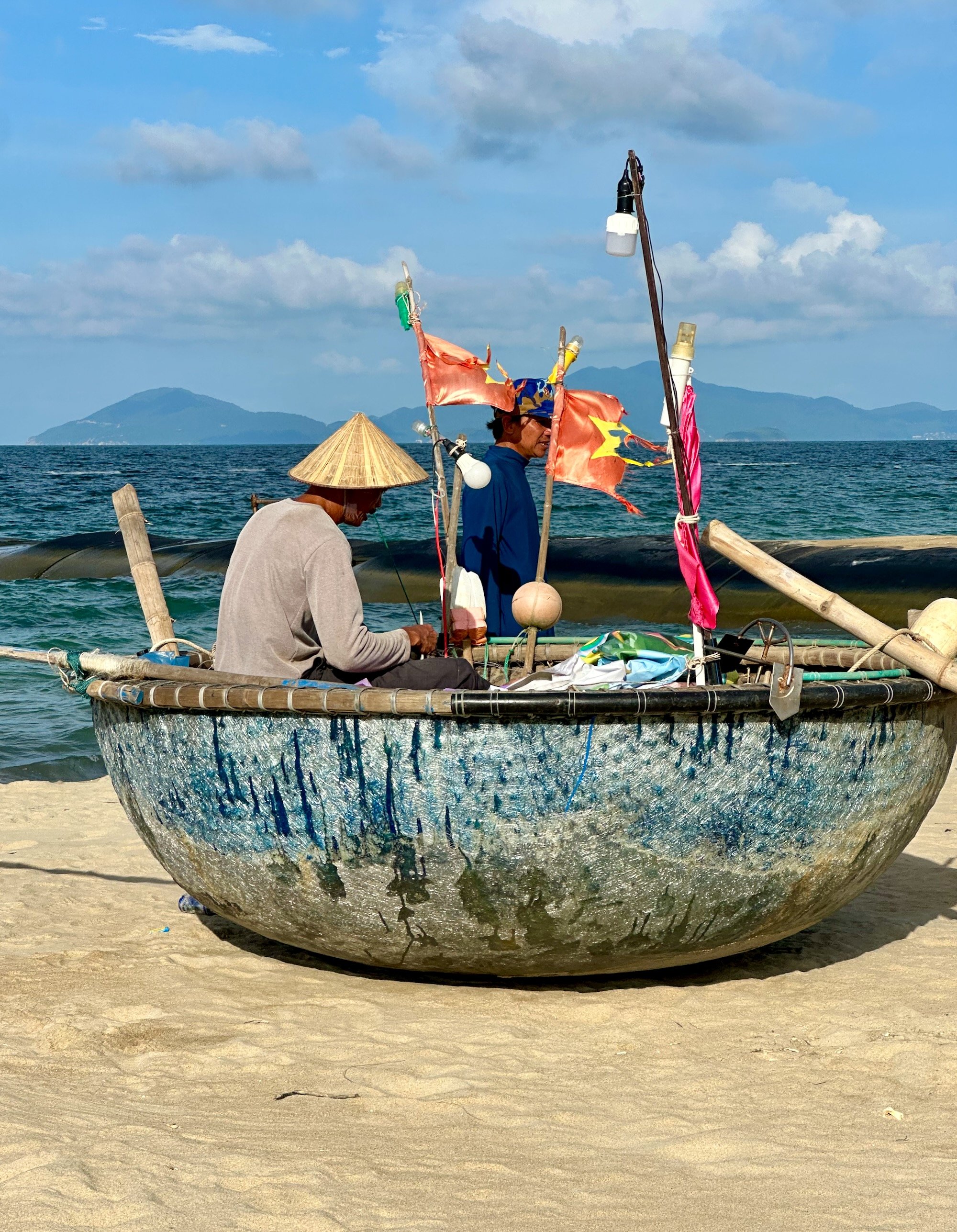 A man in a traditional Vietnamese hat sitting in a basket boat on the beach.