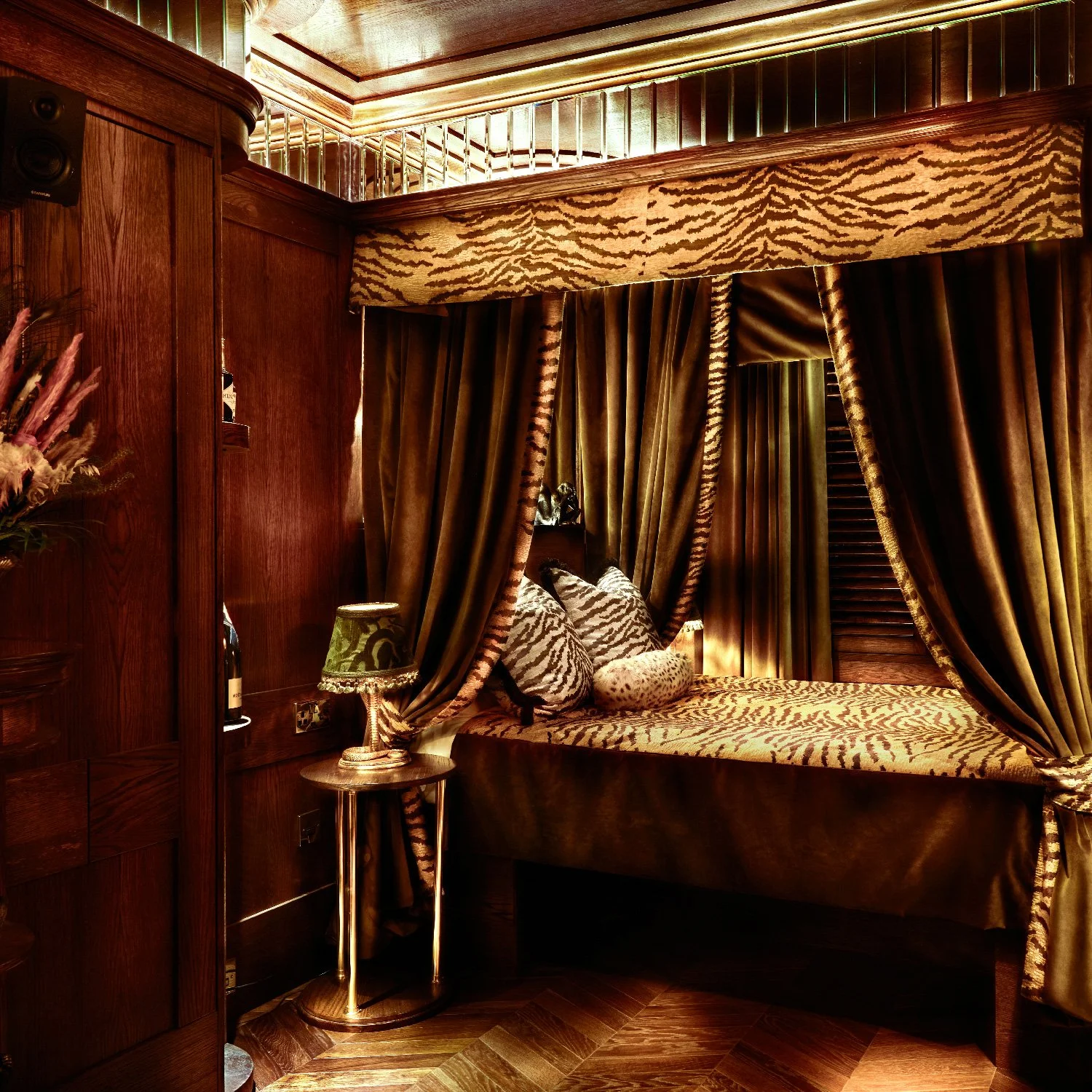 House of Gods boutique luxury hotels in Edinburgh, Glasgow & A cozy, luxurious bedroom corner with a canopy bed draped in tiger print and gold velvet curtains, a gold bedside table with a green patterned lamp, and polished wood paneling and flooring.