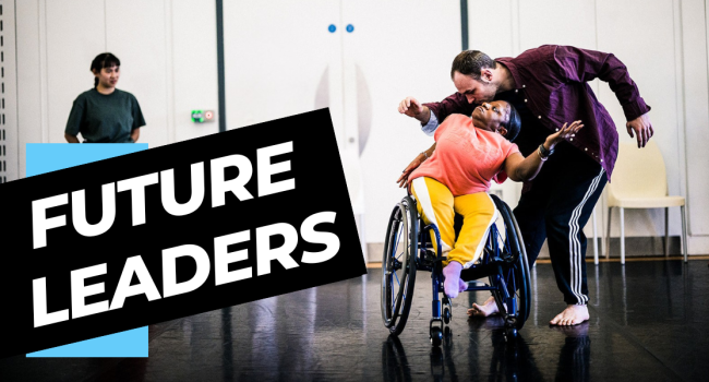 A brightly dressed dancer using a wheelchair leans back into a standing dancer who curves over her