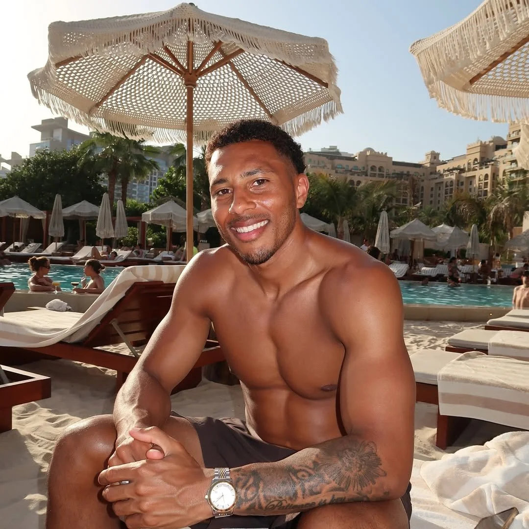 A smiling man with short hair and tattoos on his arm, sitting by a pool at a resort, wearing black shorts and a watch, with white umbrellas and lounge chairs in the background.