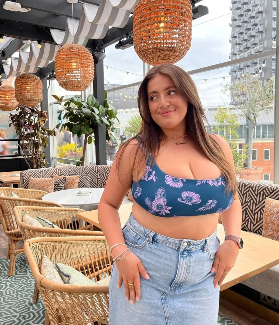 A young woman with long brown hair wearing a blue floral crop top and light blue high-waisted jeans, standing inside a cozy, well-lit cafe with bamboo hanging lamps, patterned cushions, and outdoor city scenery visible through large windows.