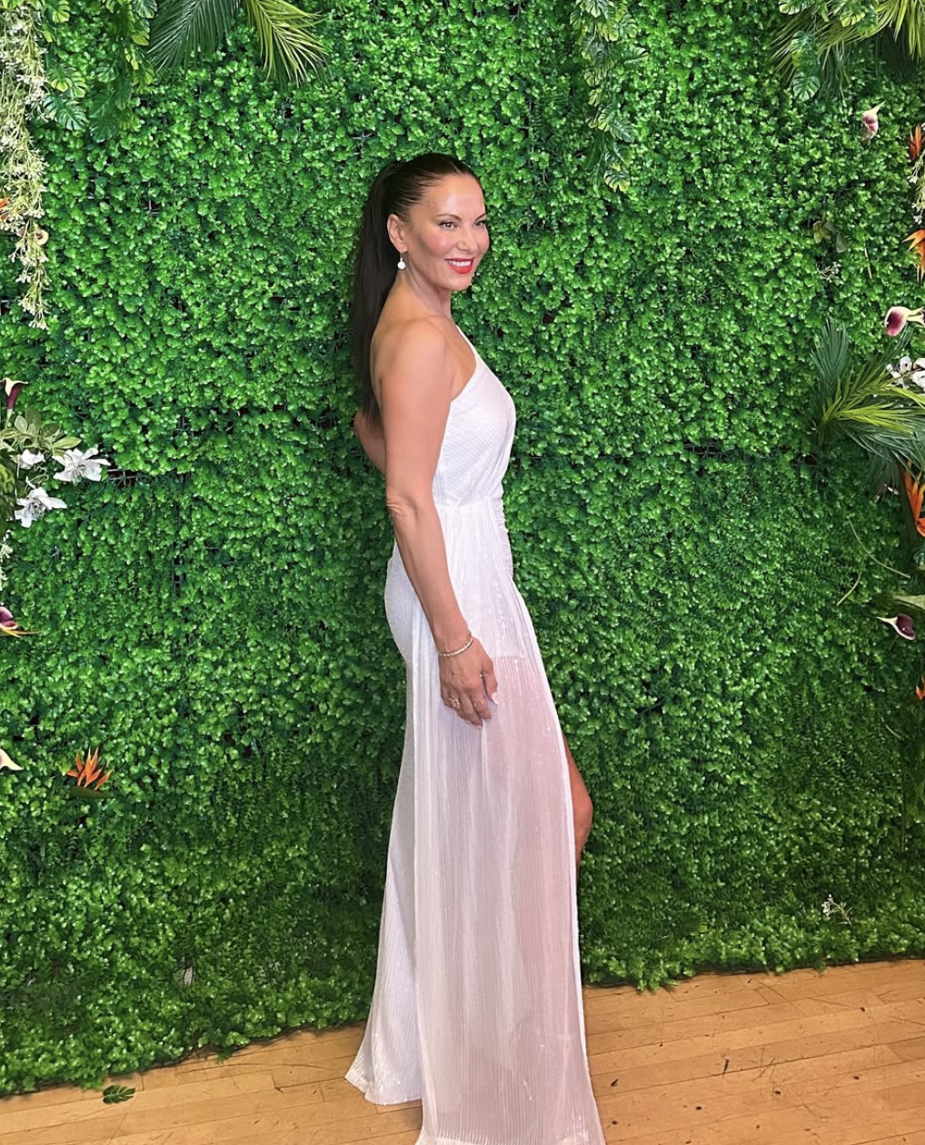 A woman in a white strapless gown standing in front of a lush green leafy wall with plants and flowers, smiling at the camera.