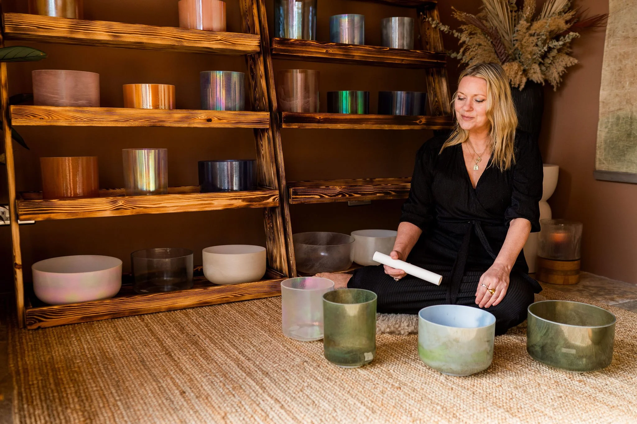 A woman sitting cross-legged on a beige carpet playing crystal singing bowls. She is holding a mallet in her right hand and is surrounded by several glass bowls of various sizes and colors. The background features wooden shelves with more bowls and a decorative dried plant arrangement.