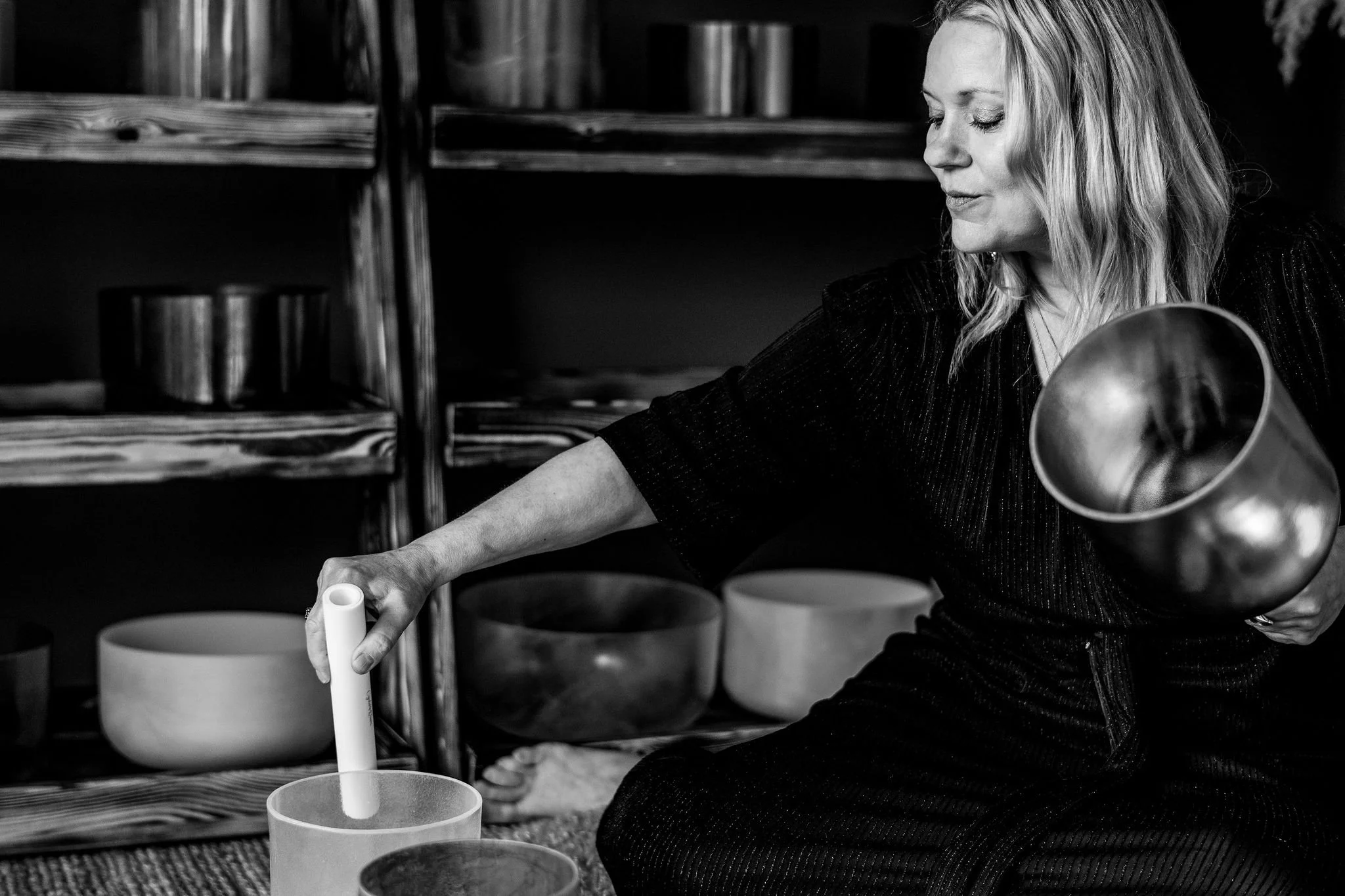 A woman with blonde hair in a black dress, sitting and playing crystal singing bowls in a room with wooden shelves containing more bowls.