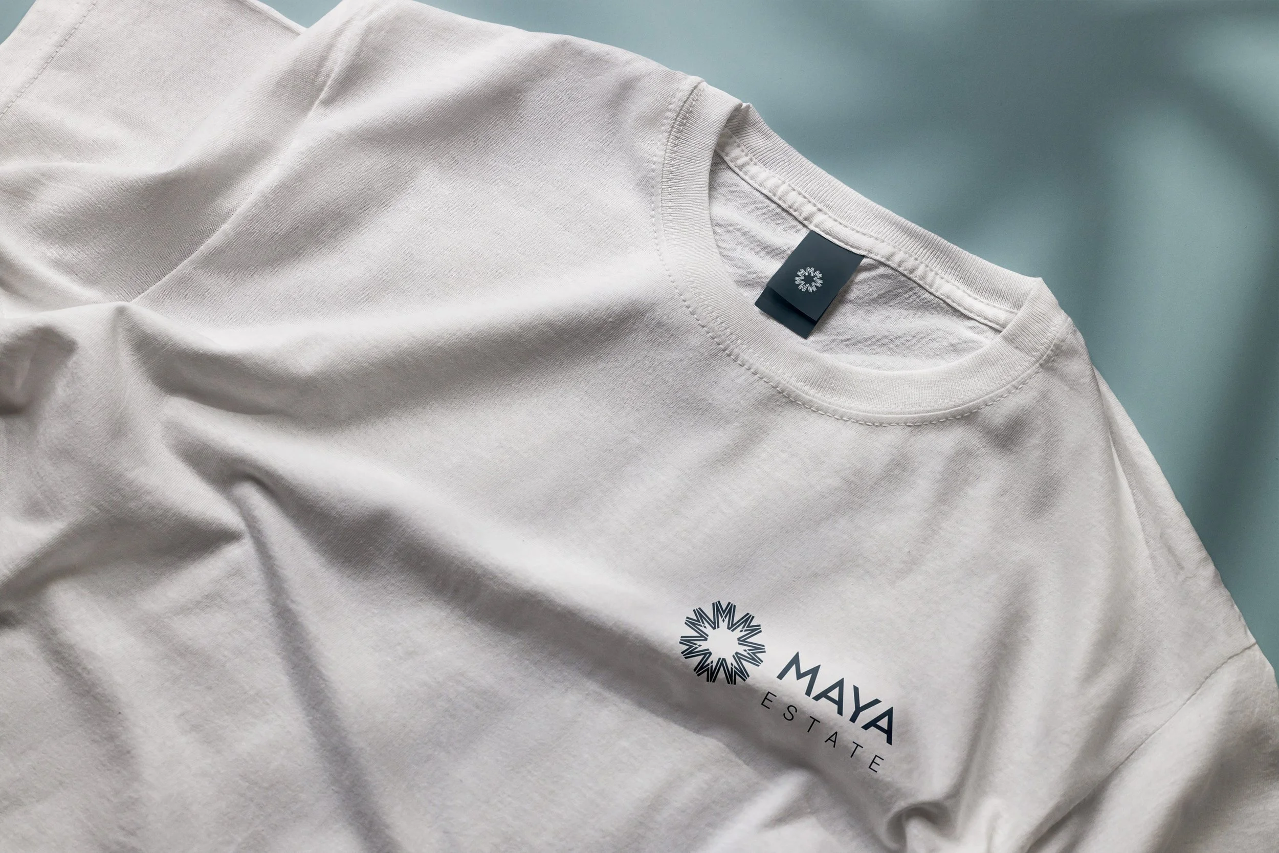 White T-shirt with 'Maya Estate' logo on fabric, laid on a pale blue surface.
