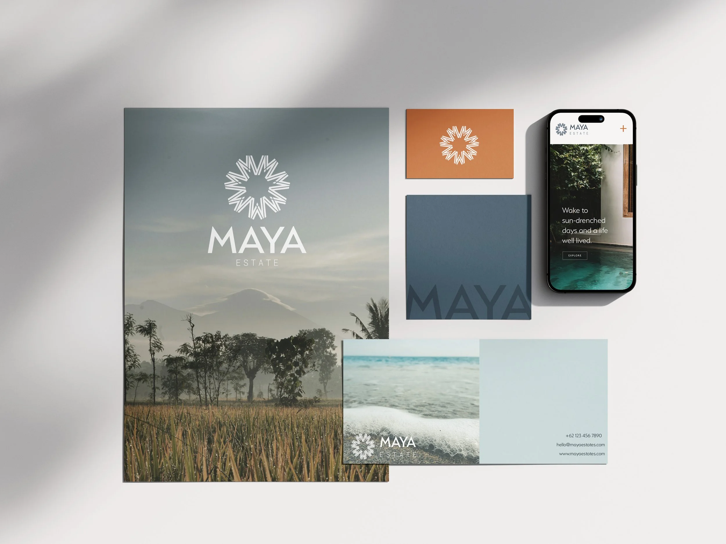 Collection of branding materials for Maya Estate, including a large landscape poster with mountains and trees, a smaller beach photo, a navy blue and orange color swatch, and a smartphone displaying the Maya Estate website with poolside image and tex