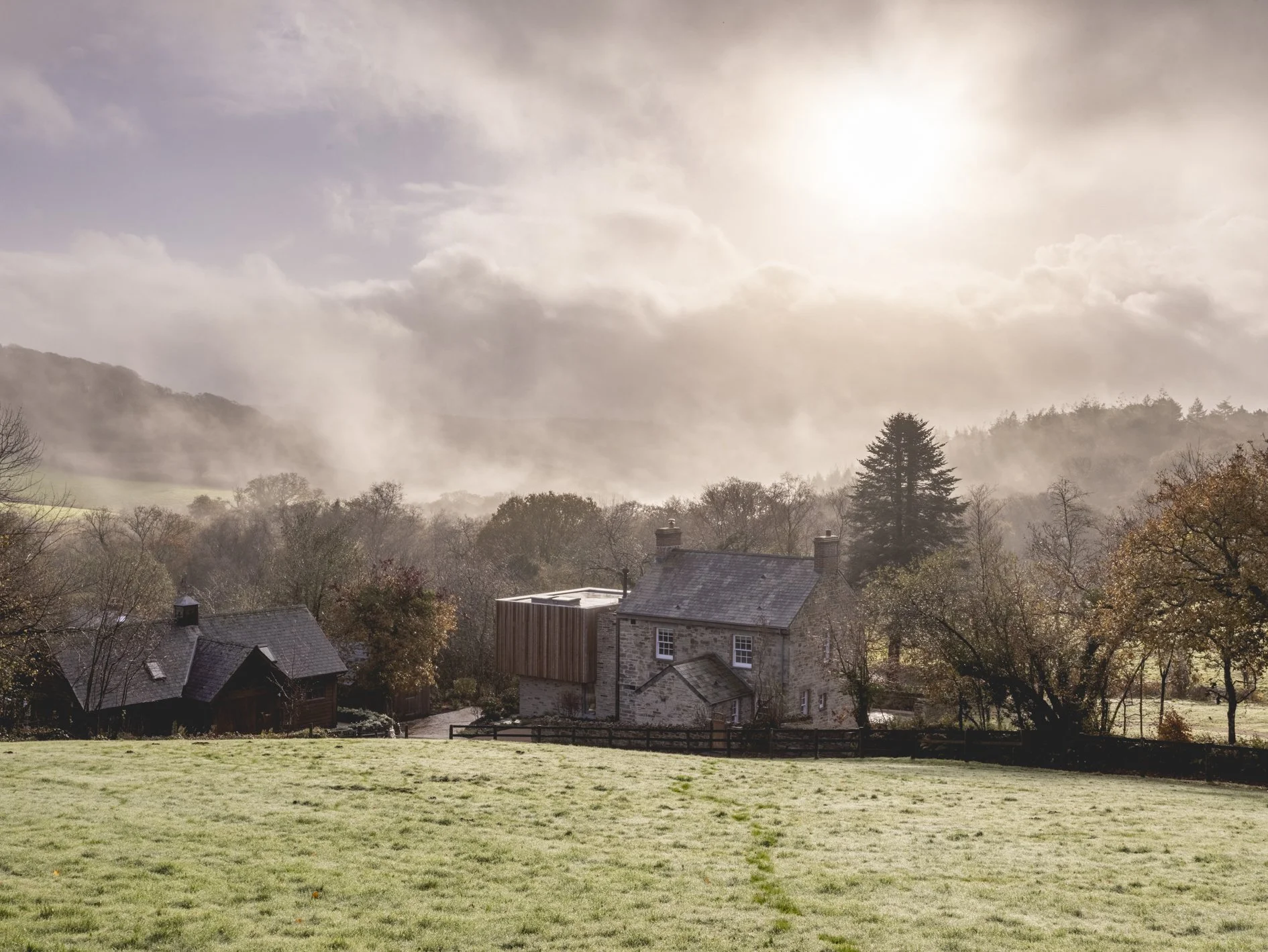 A rural landscape with a grassy field in the foreground, stone houses with chimneys, trees with autumn foliage, and fog-covered hills under a cloudy sky with sunlight.