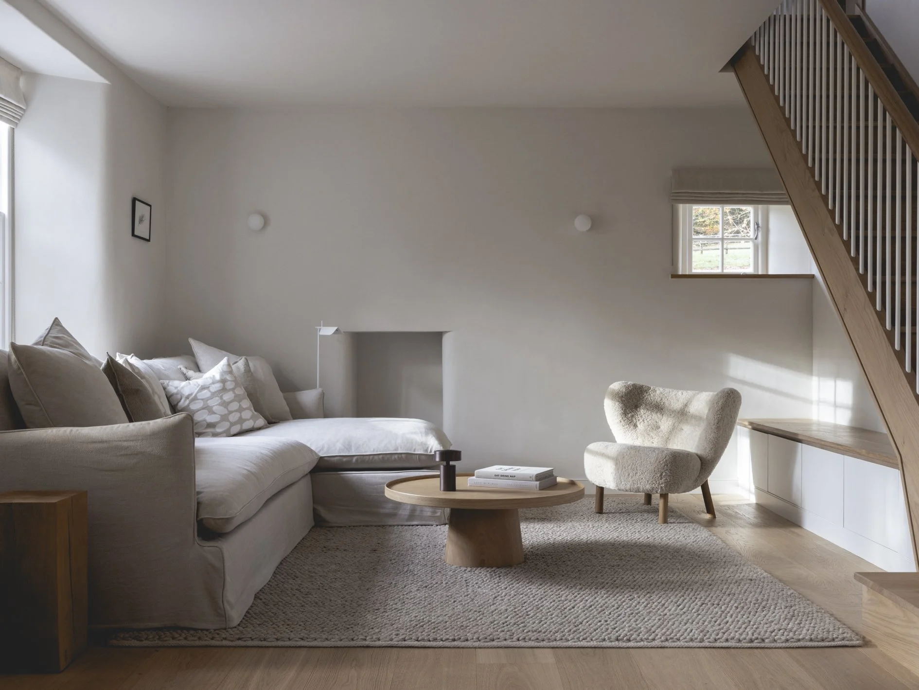 A cozy living room with a beige sectional sofa and a plush armchair, a round wooden coffee table with books, and large windows allowing natural light, under a wooden staircase.