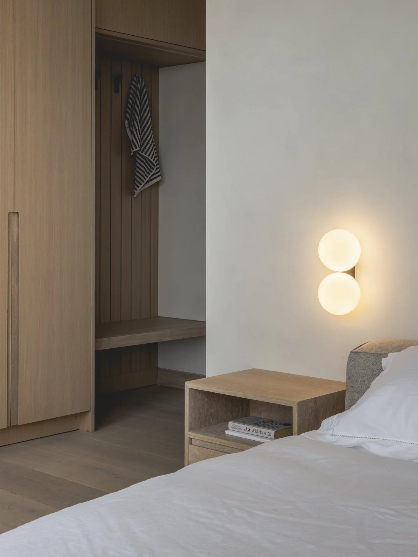 A bedroom with a wooden nightstand, a bed with white sheets, wall-mounted round lights, and an open closet with a striped towel hanging inside.