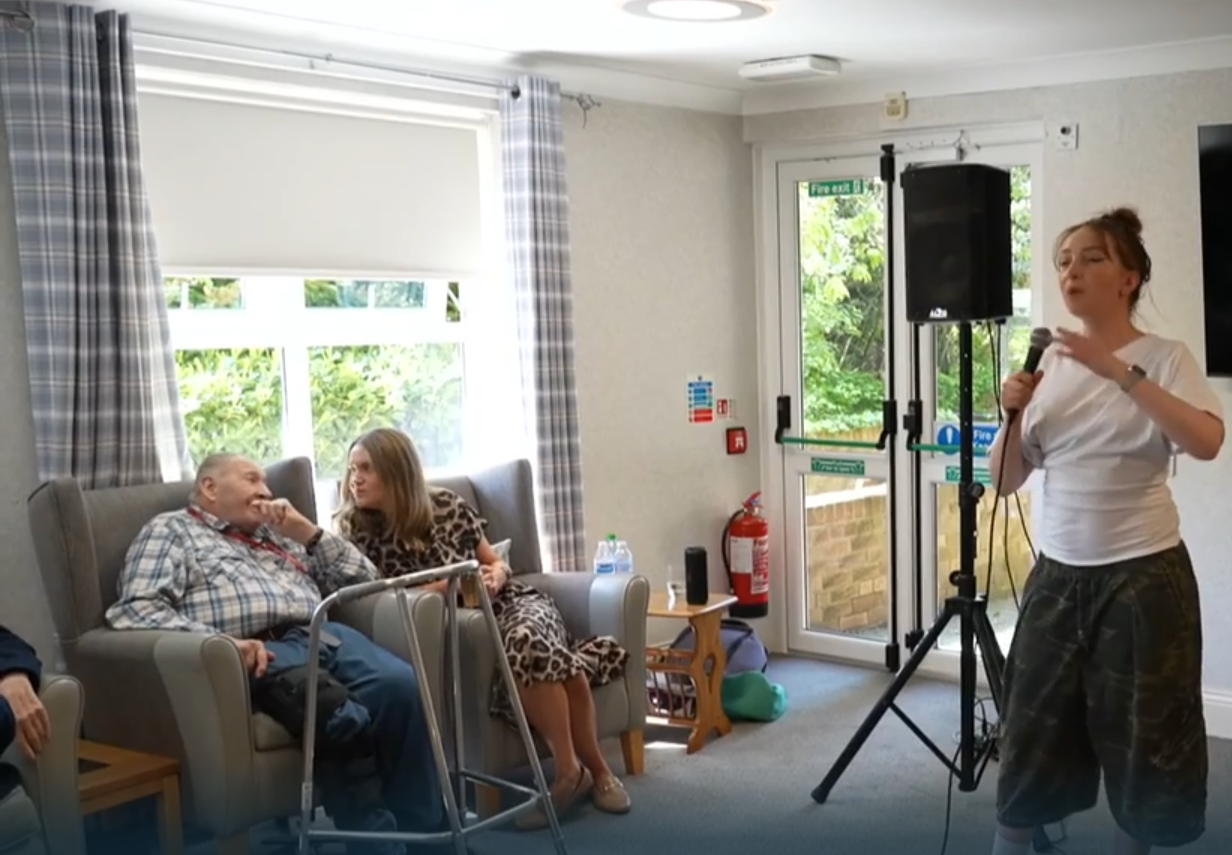 A woman singing into a microphone in front of an audience of elderly people in a room with a door and window showing greenery outside.