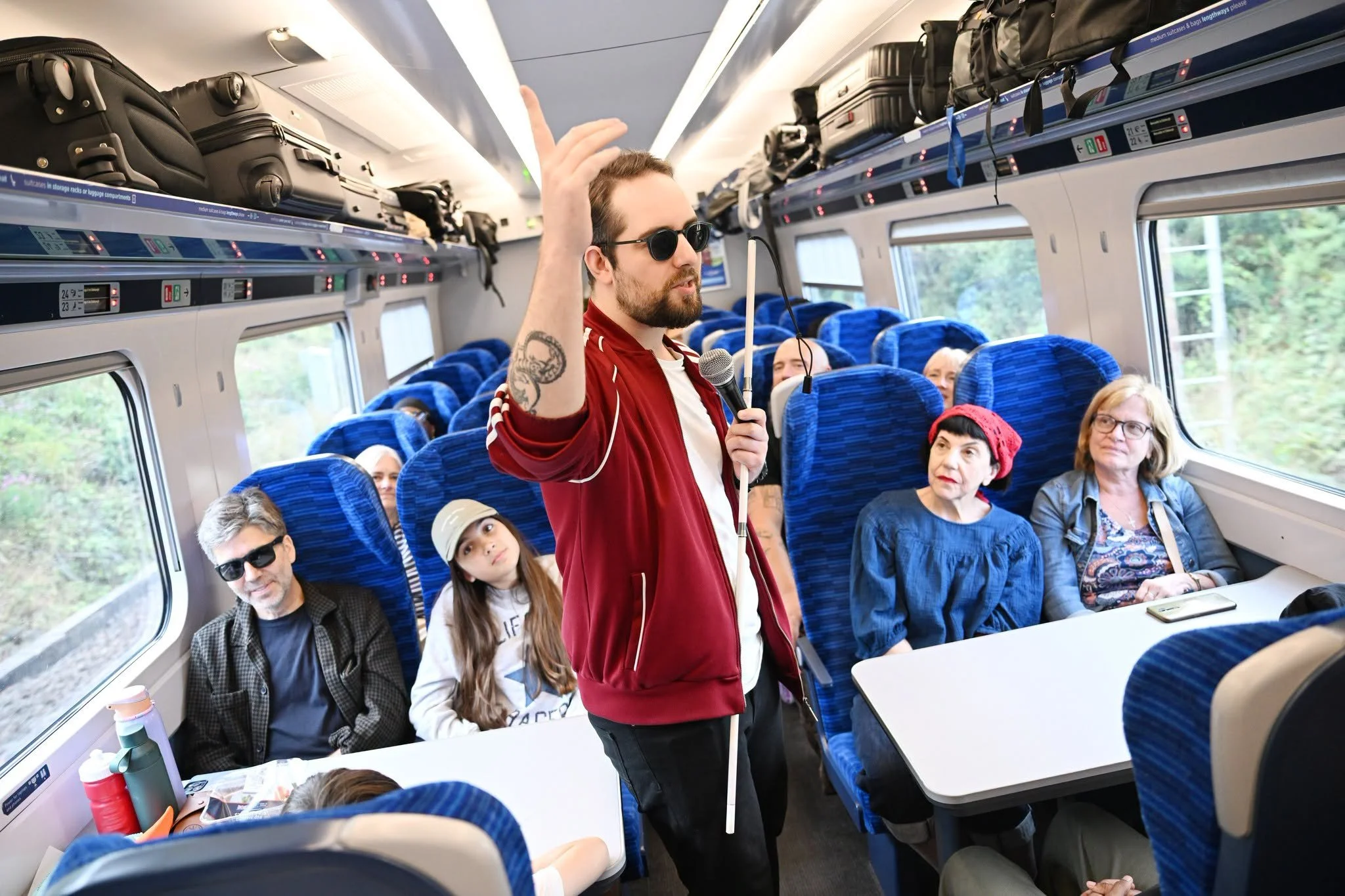 A man with dark sunglasses and a beard, wearing a red jacket, holding a microphone and raising his hand, speaking to a group of seated passengers on a train. The passengers, of various ages, are listening, with some looking at the man and others engaging in different directions. The train interior has blue seats, large windows showing greenery outside, and overhead luggage compartments filled with suitcases and bags.