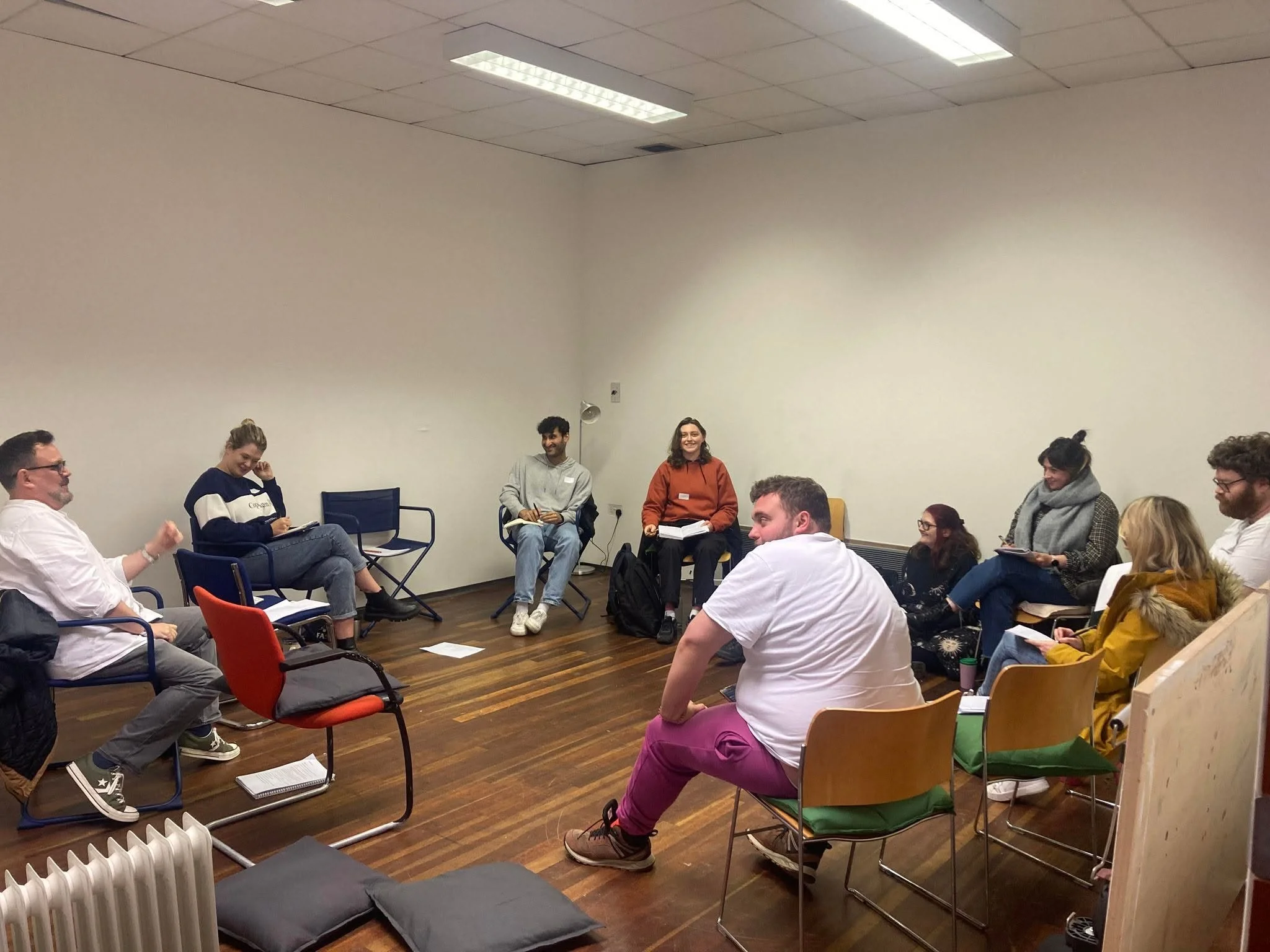Group of people seated in a circle participating in a discussion or workshop in a room with white walls and hardwood floors.
