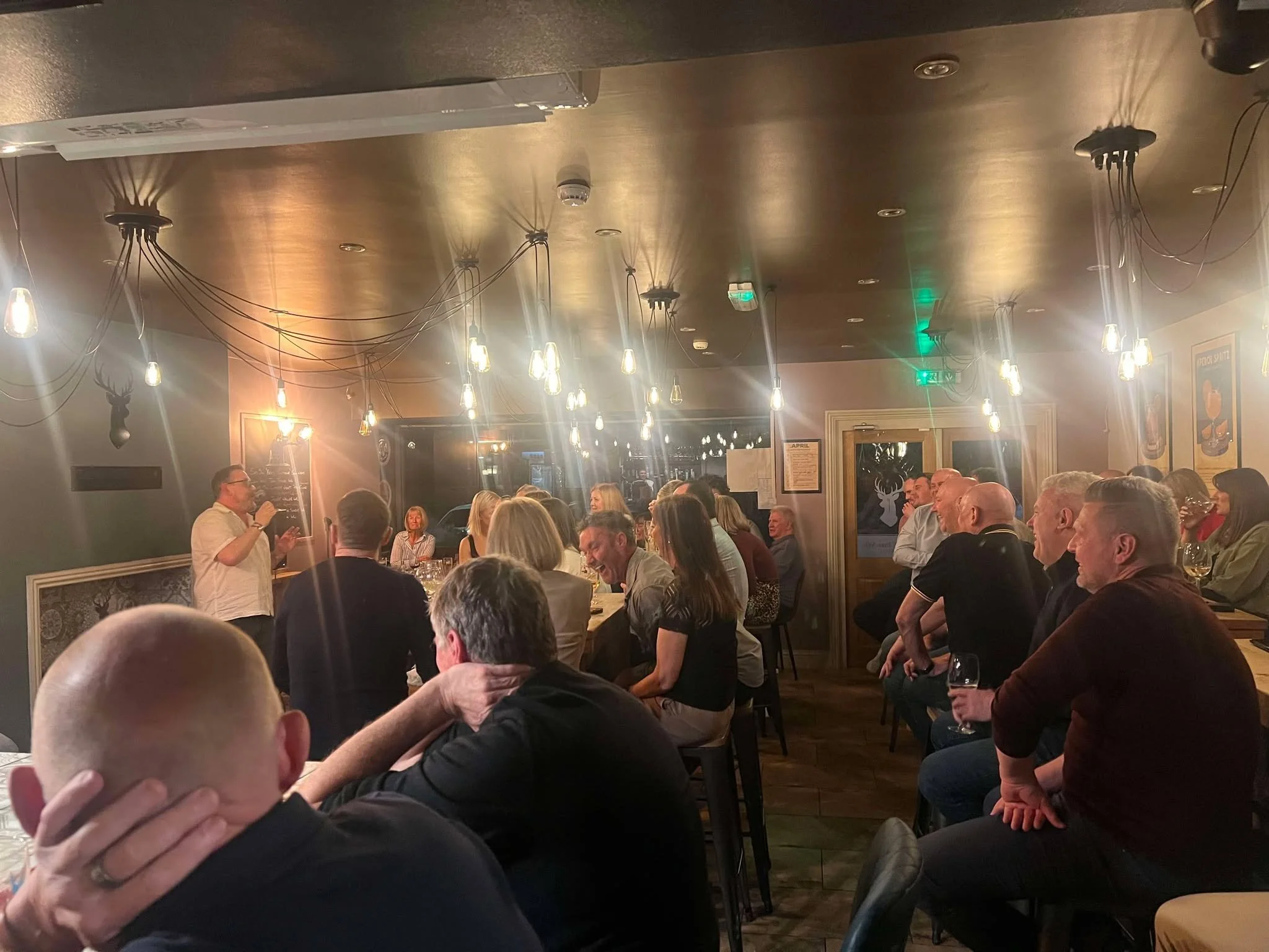 Group of people seated and standing in a warmly lit restaurant or pub with wooden walls and industrial-style light fixtures hanging from the ceiling, engaged in conversation and enjoying drinks.