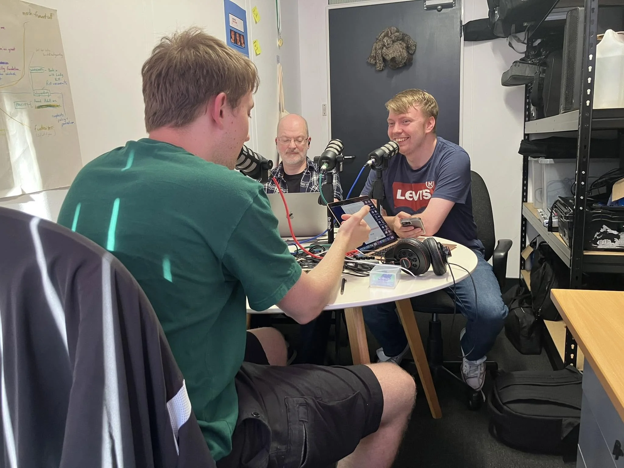 Three men sitting at a table, engaged in a discussion or podcast recording, with microphones and recording equipment. Two of the men are looking at a phone, and the third man is listening with closed eyes. A whiteboard and shelving units with equipment are in the background.