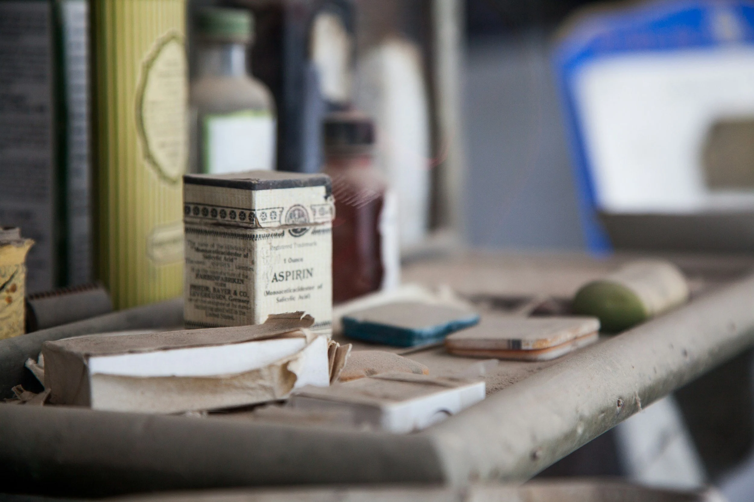 An image depicting a wooden tray with old paper and tin boxes covered in dust with a box of aspirin figuring prominently. The background is blurred but we can see that the tray is sitting in front of a bookshelf.