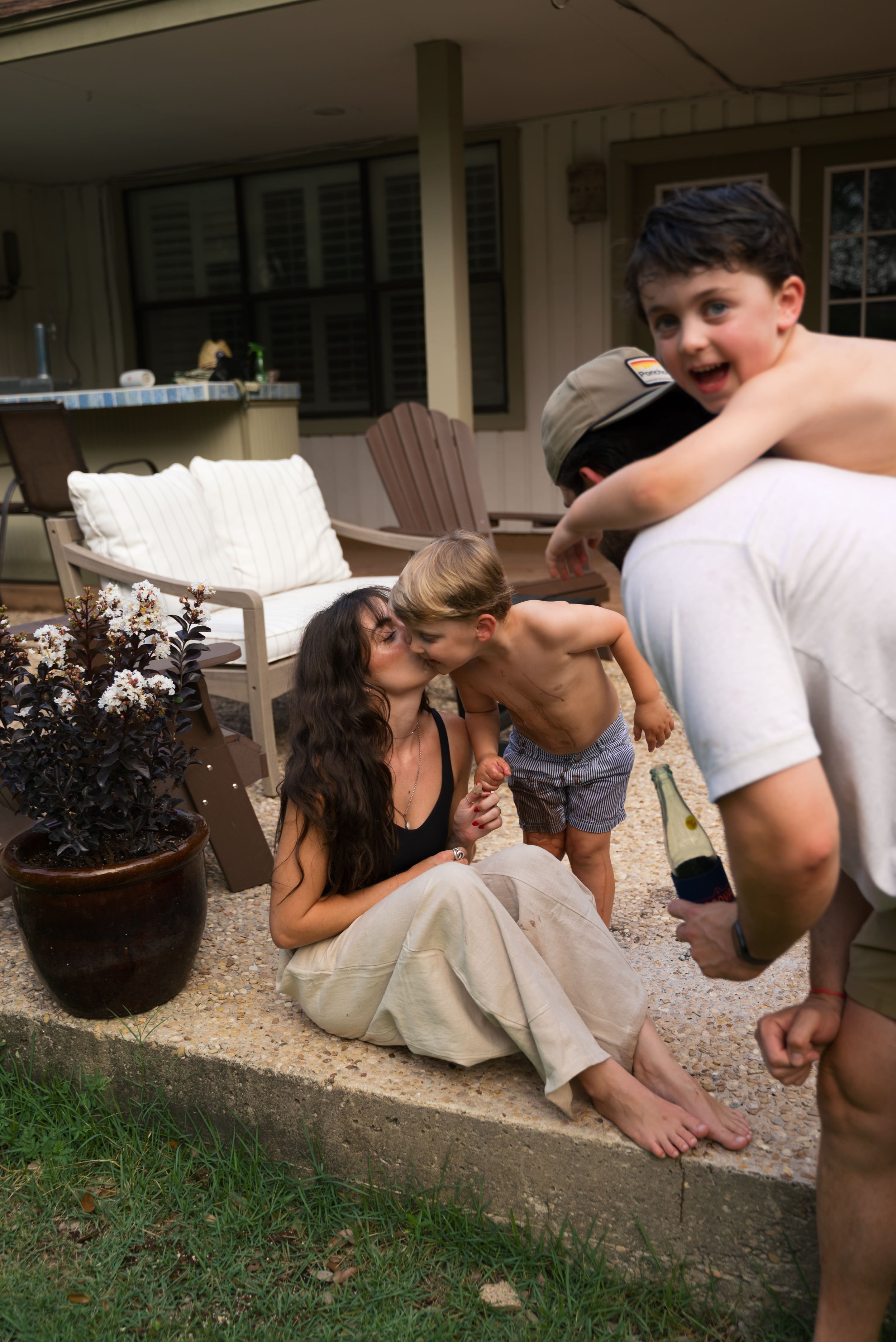 A woman sitting on the ground outside, kissing a young boy standing next to her. Two other boys are close by; one is being held on an adult's back and the other is smiling at the camera, holding a bottle. The scene is in a backyard with outdoor furniture and plants.