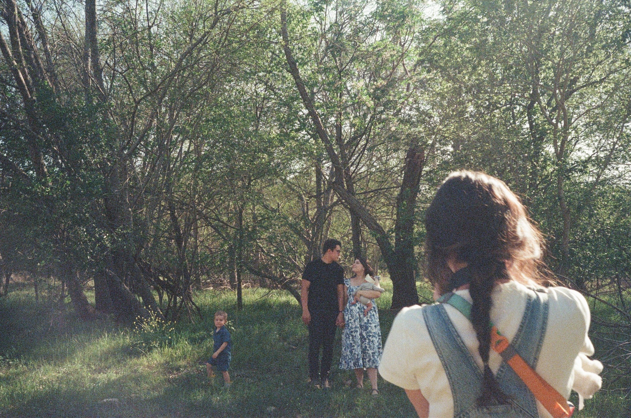 A group of four people, including a woman holding a dog, standing in a forested area with tall trees and green grass. A young boy is standing nearby, and a person with long hair and overalls is in the foreground with their back to the camera.
