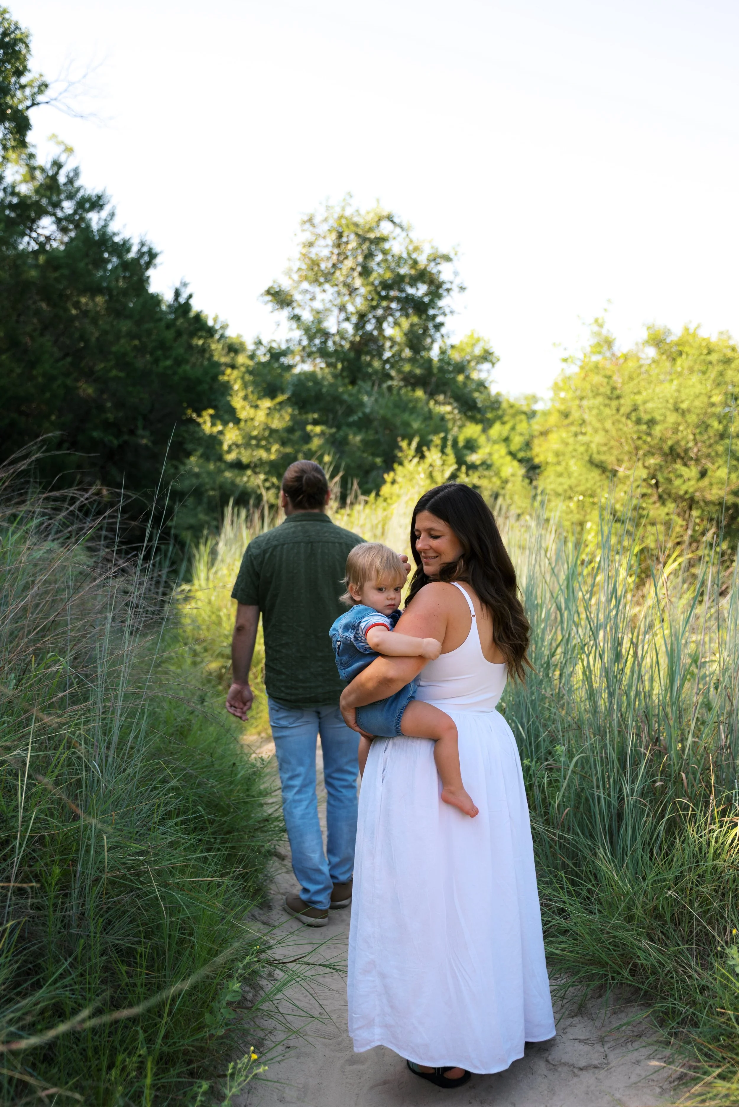 A woman with long dark hair carrying a young child in a white dress on a nature trail, with a man walking ahead in a green shirt and light blue jeans among tall grass and trees in Madison, Wisconsin