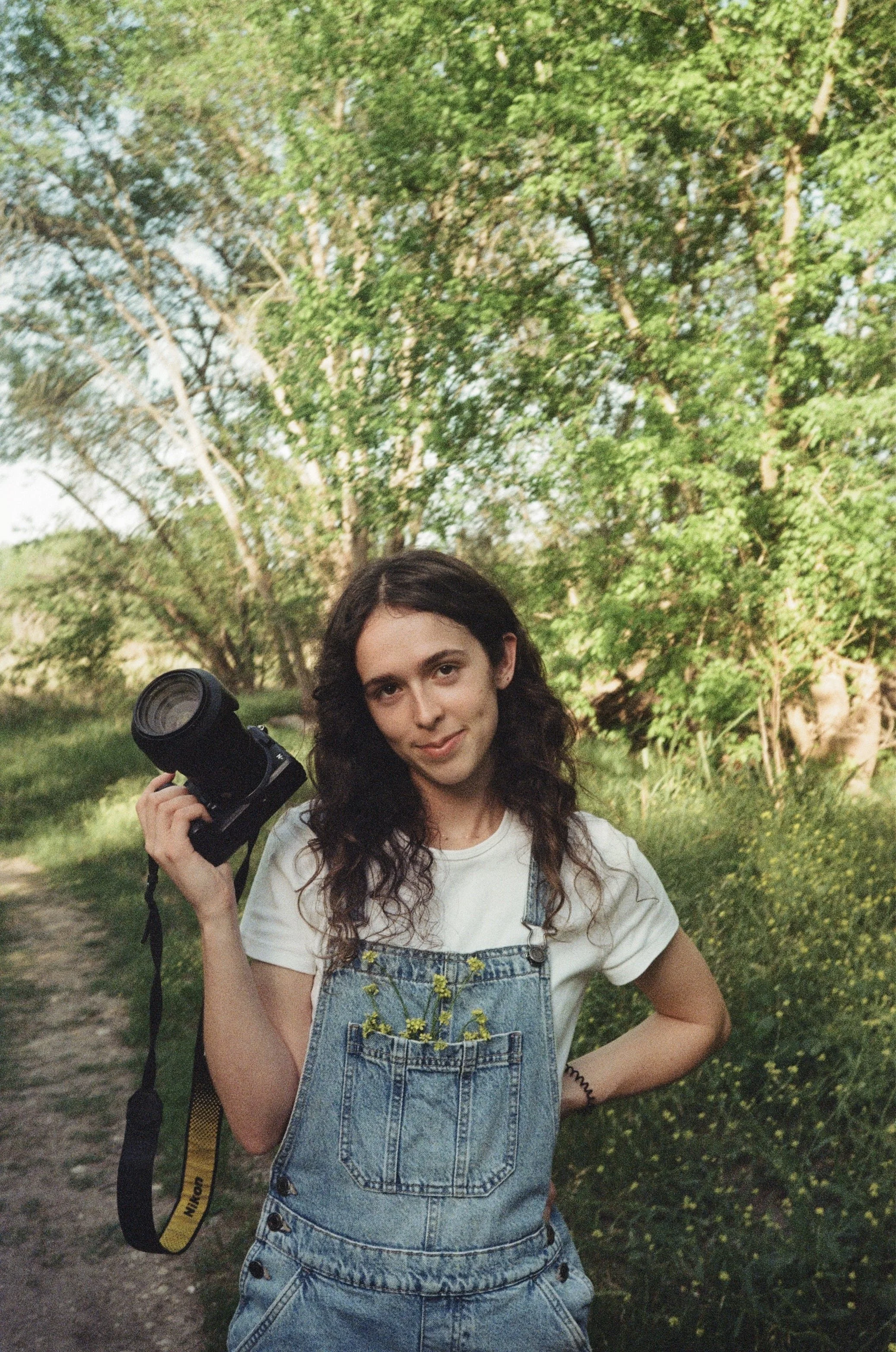 A young woman with long dark curly hair wearing a white t-shirt and denim overalls standing outdoors on a trail surrounded by green trees and grass, holding a camera in her right hand and looking at the camera.