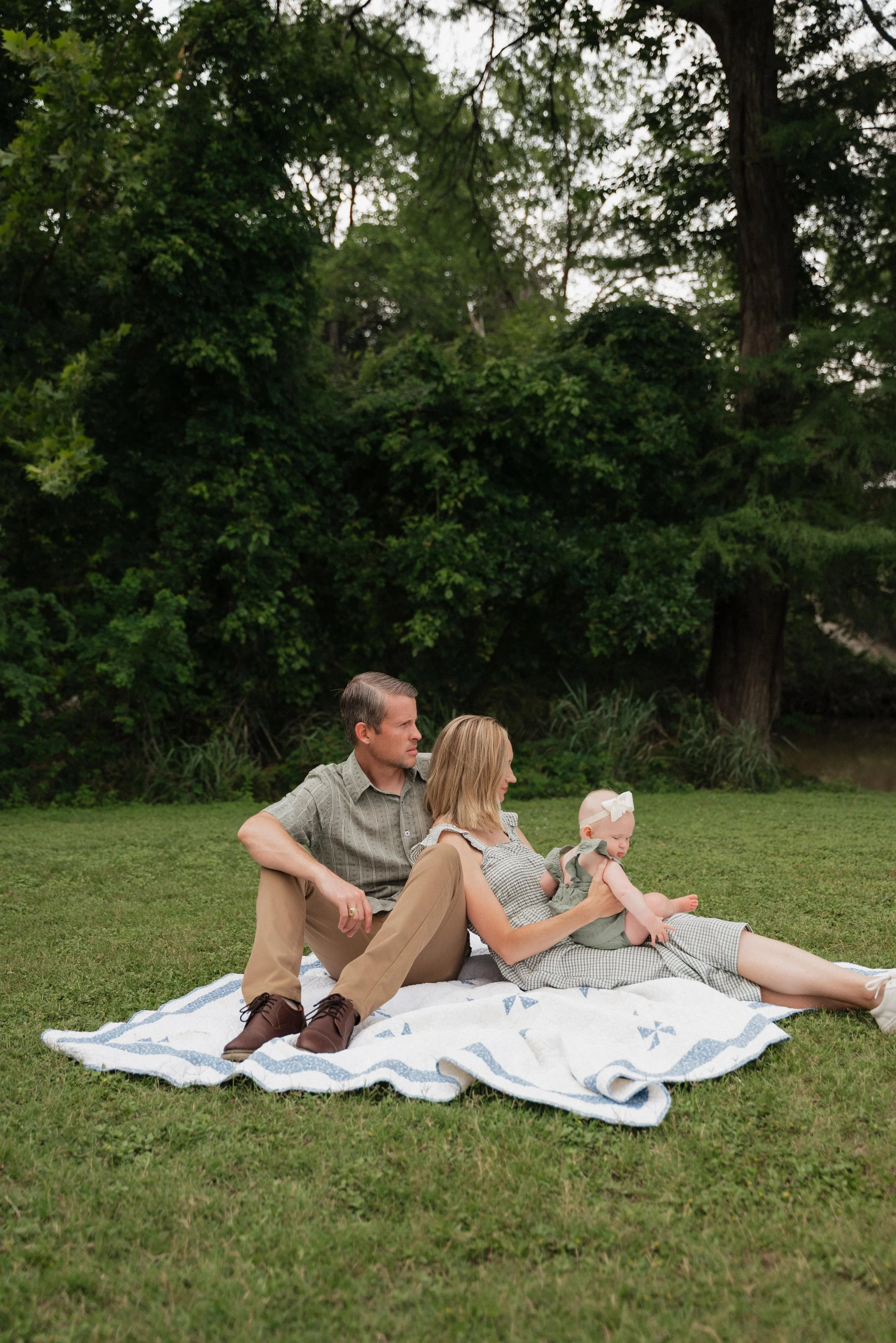 A family of three sitting on a blanket outdoors near a wooded area, with the mother holding a baby girl, the father sitting beside them, all enjoying a peaceful moment.