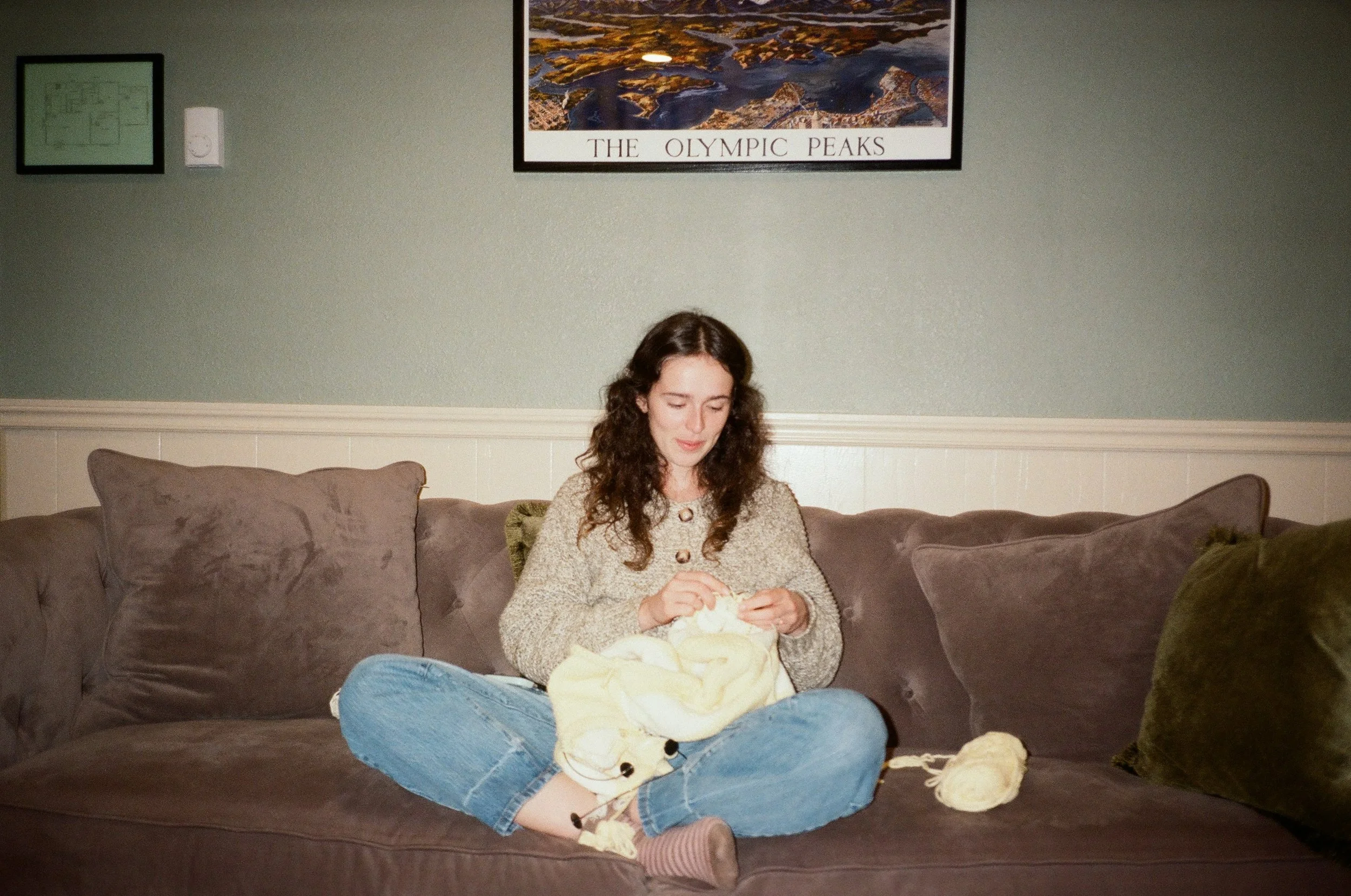 A woman with dark curly hair sitting cross-legged on a beige couch, knitting with yellow yarn. She is wearing a beige sweater and blue jeans, with a yarn ball to her right. The background includes a wall with framed pictures and a large poster of the Olympic Peaks.
