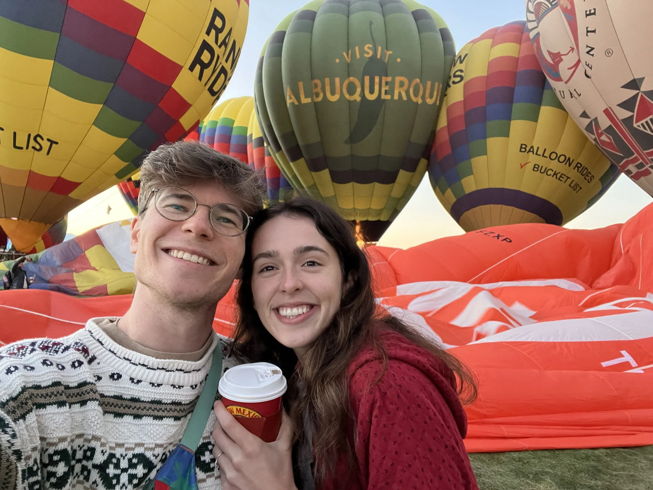 Two smiling people taking a selfie at a hot air balloon festival, with multiple colorful hot air balloons in the background.