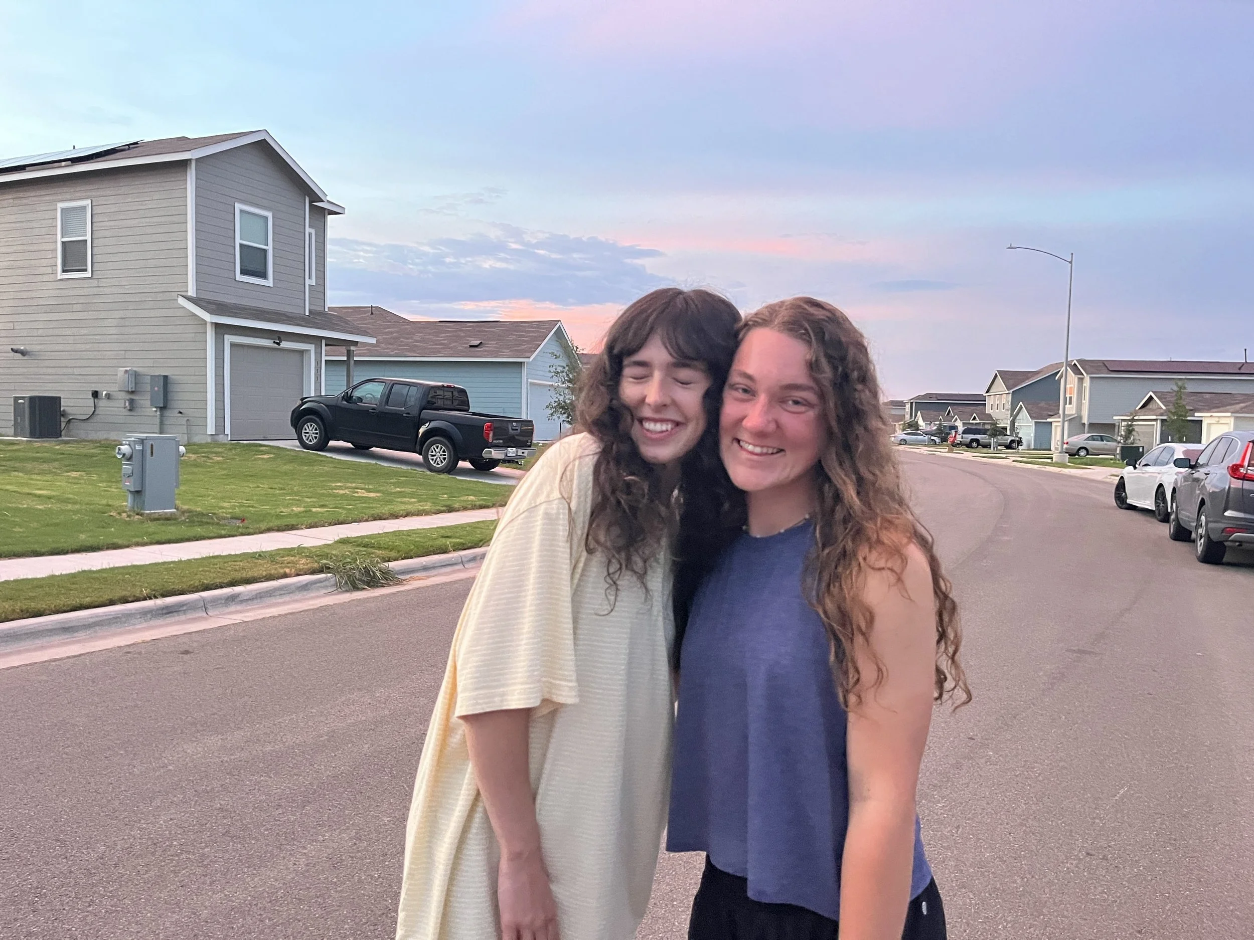Two young women with curly hair smiling and hugging on a suburban street during sunset.