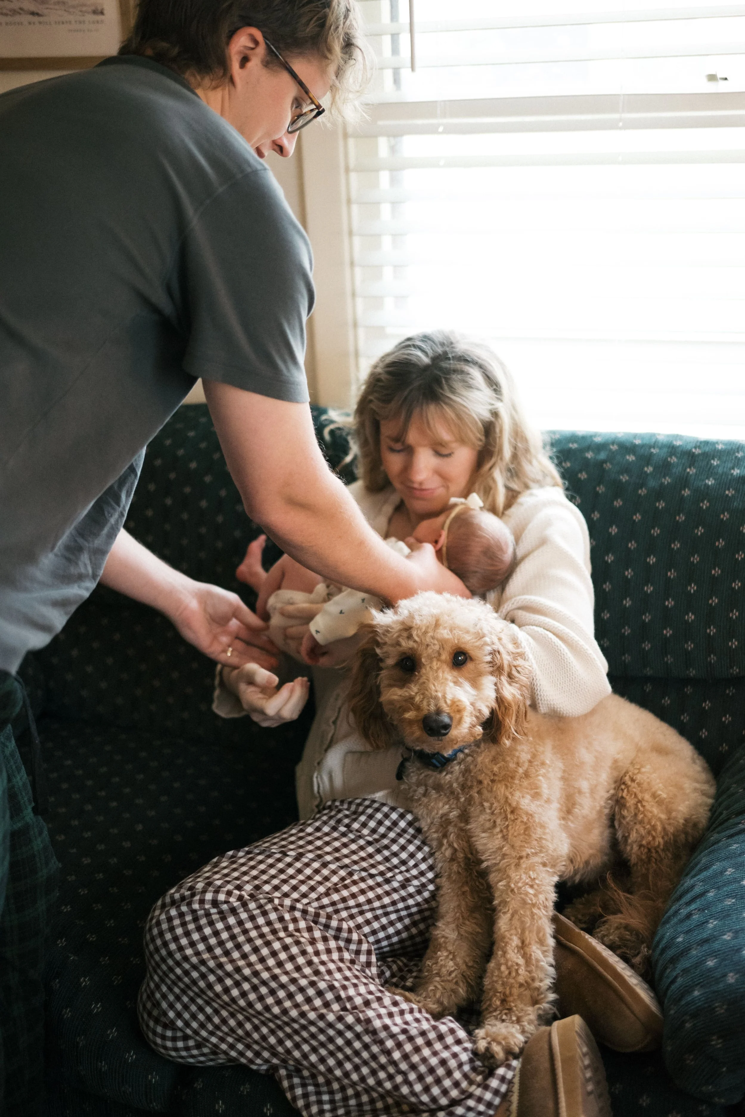 A woman holding a newborn baby with an older woman and a small dog sitting on a couch near a window.