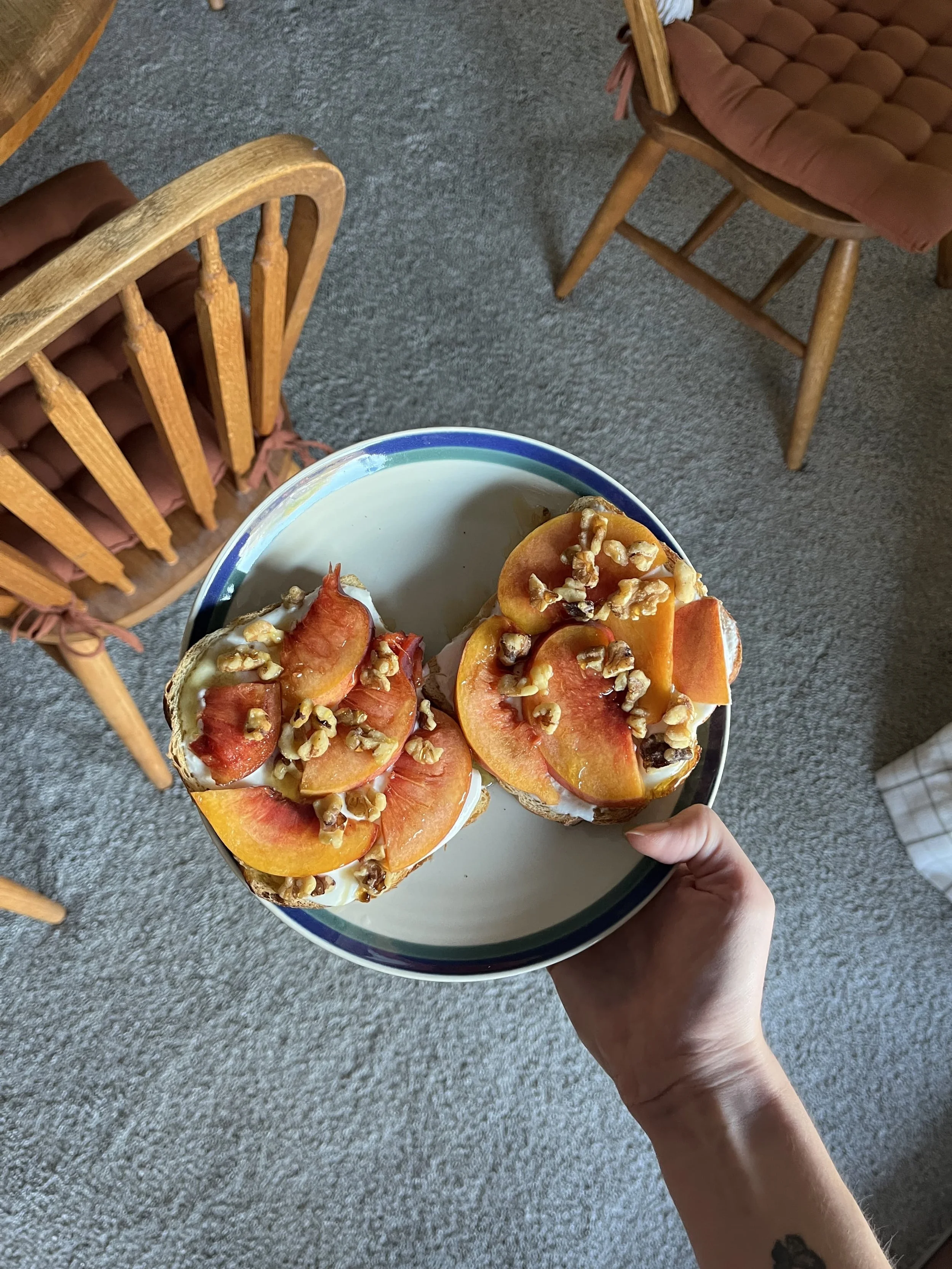A person holding a plate with two slices of bread topped with yogurt, peach slices, and chopped nuts, with a background of wooden chairs and a carpeted floor.