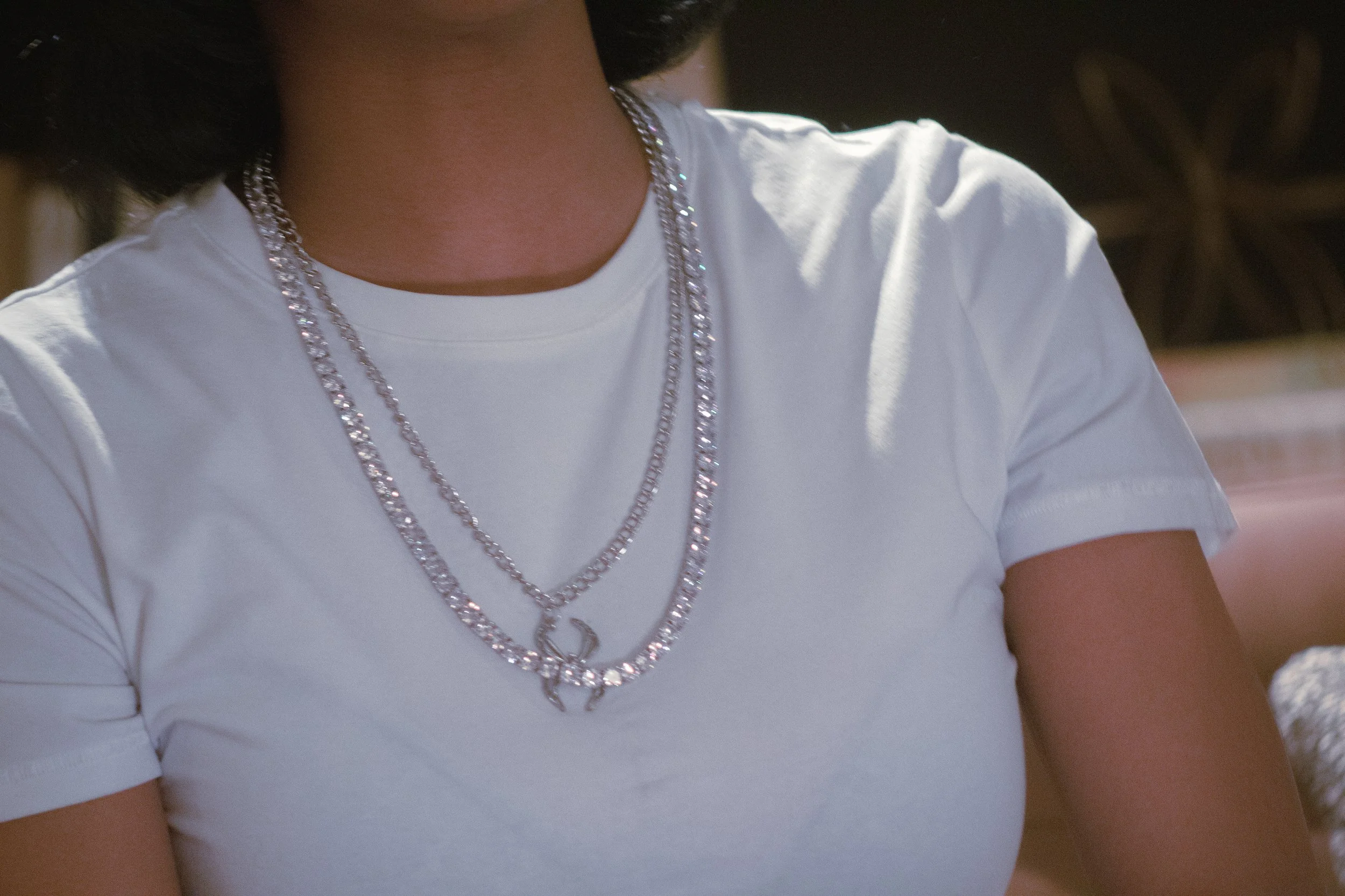 Close-up of a person's neck and upper chest wearing layered silver necklaces, dressed in a white t-shirt.