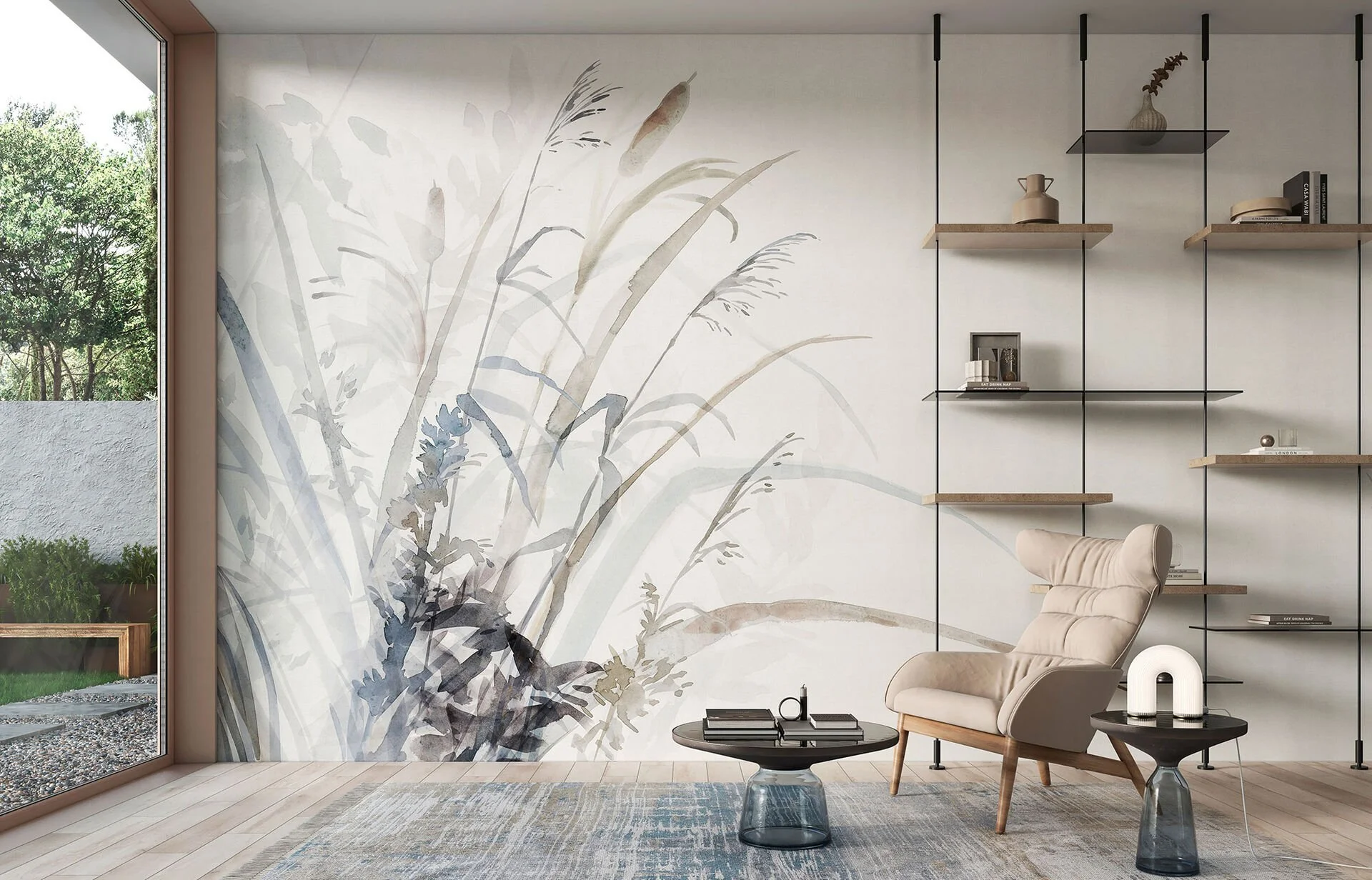 Modern living room with a large watercolor botanical mural on the wall, a beige armchair with a wooden base, a round glass-top coffee table with books and candles, a smaller black side table with a decorative lamp, and wall-mounted wooden shelves with decor items. Large window shows a garden outside.