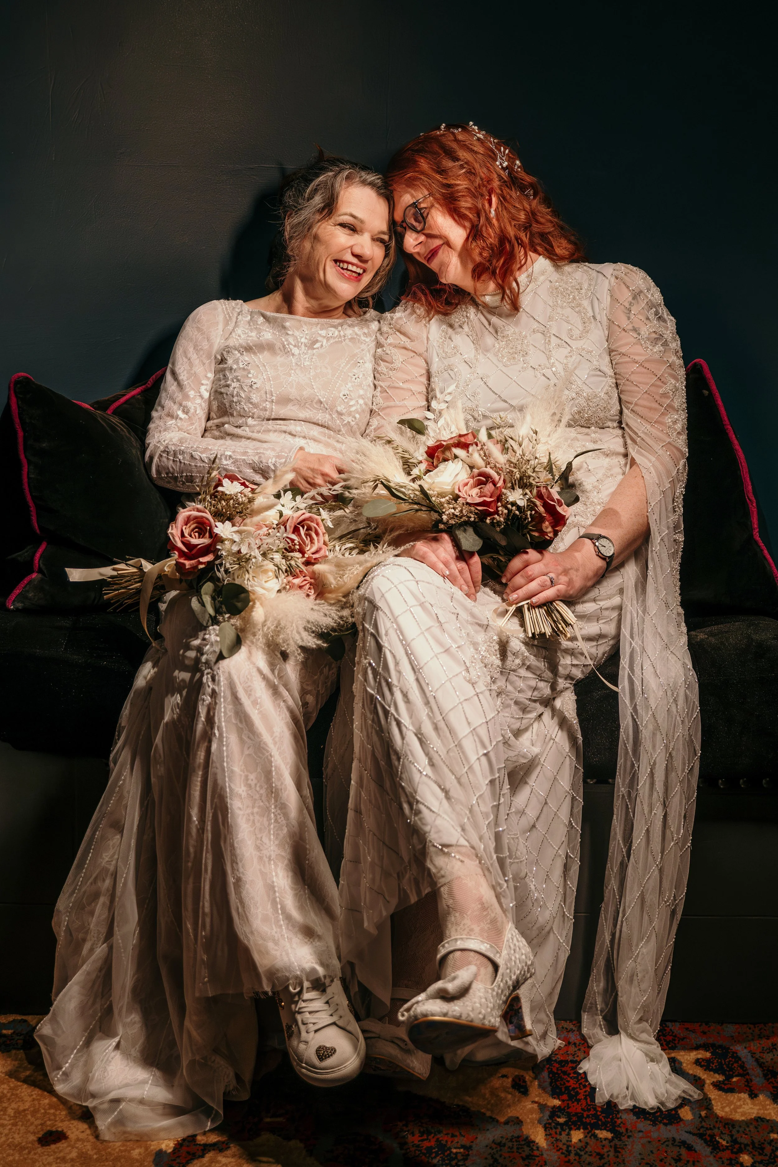 2 brides sit with their temples touching and they are holding bouquets and smiling.