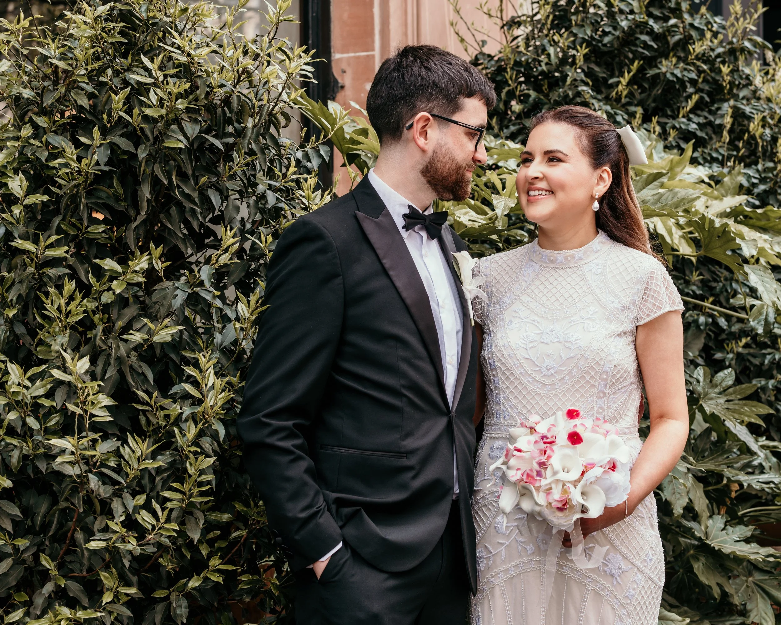 A bride and groom dressed in wedding attire standing close together outdoors, holding a bouquet of white and pink flowers, with green foliage in the background.