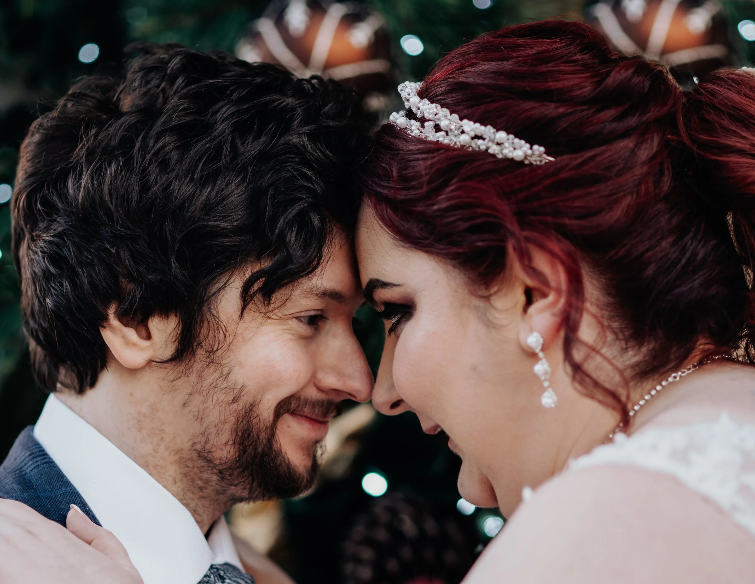 Close-up of a man and woman touching foreheads, smiling, with wedding attire and jewelry, in a festive setting with blurred lights in the background.