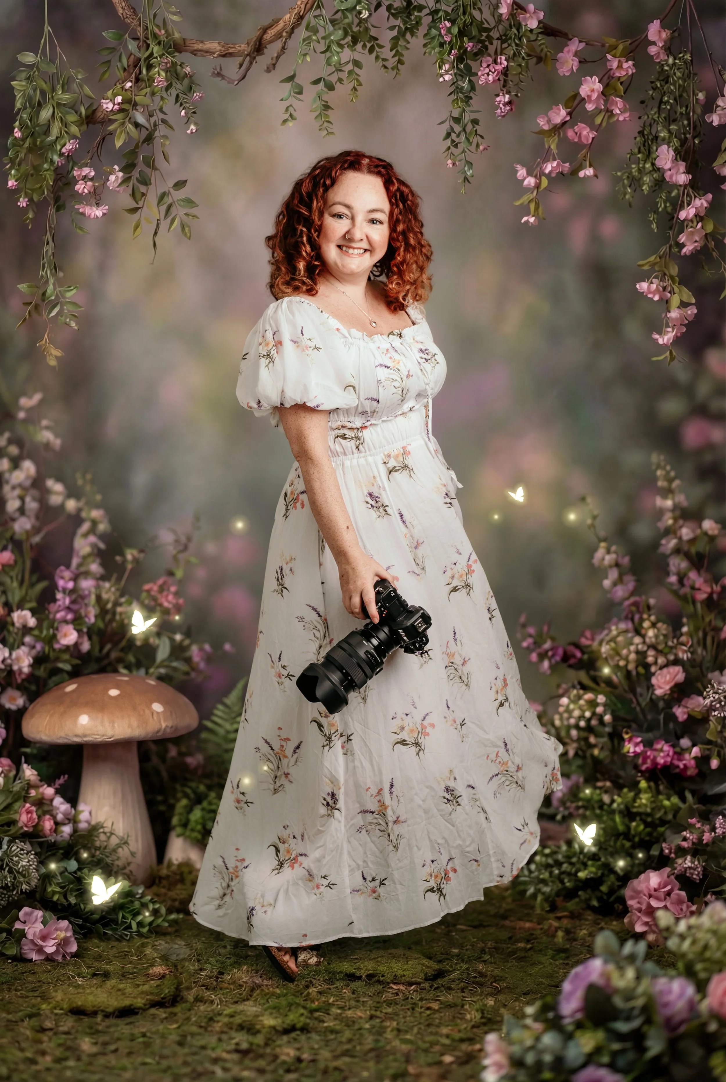 A woman with curly red hair wearing a white floral dress holding a camera in a garden decorated with pink flowers, ferns, mushrooms, and fairy lights.