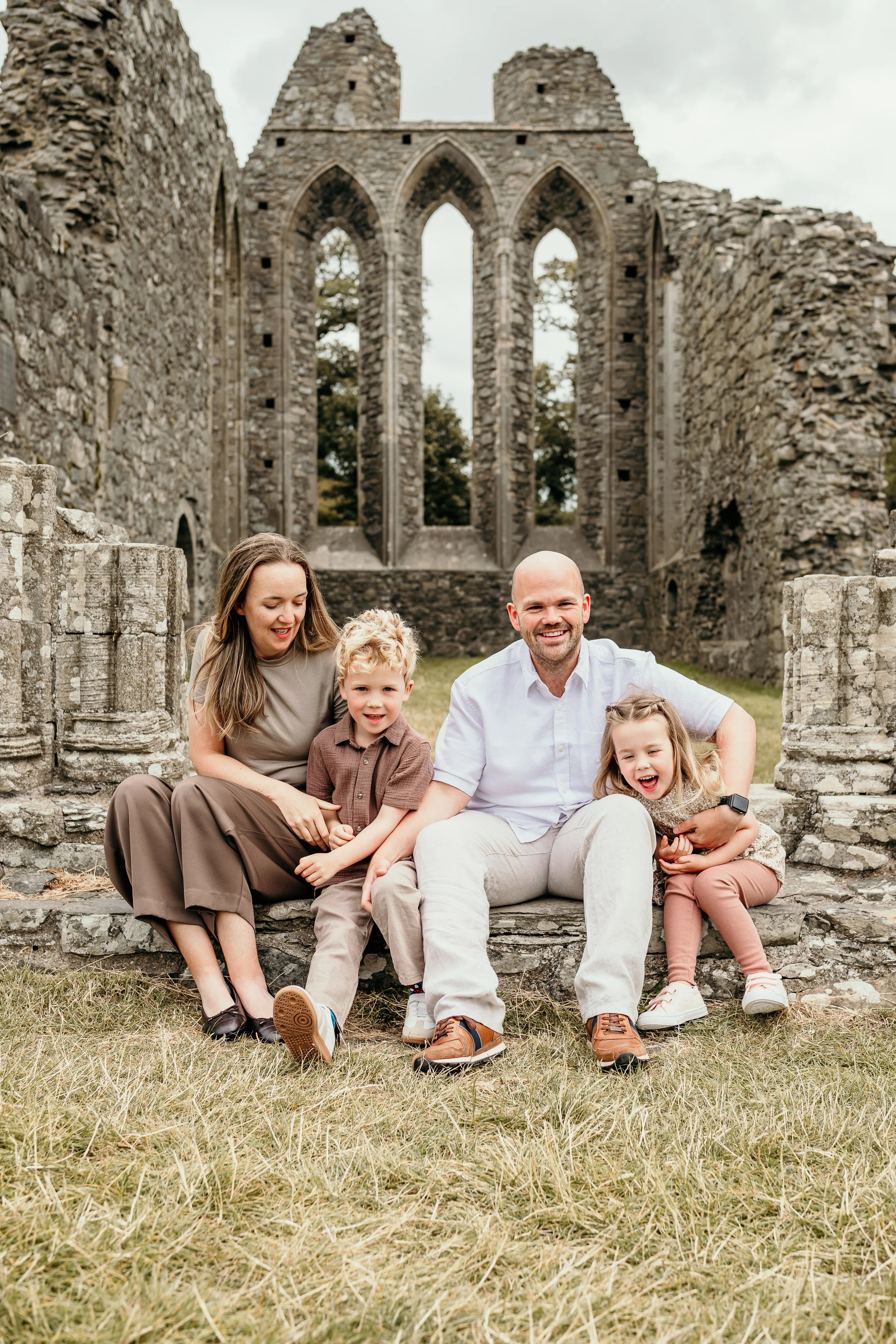 Family of four sitting in the ruins of Inch Abbey: mum, dad, young boy, young girl. Young girl is laughing.