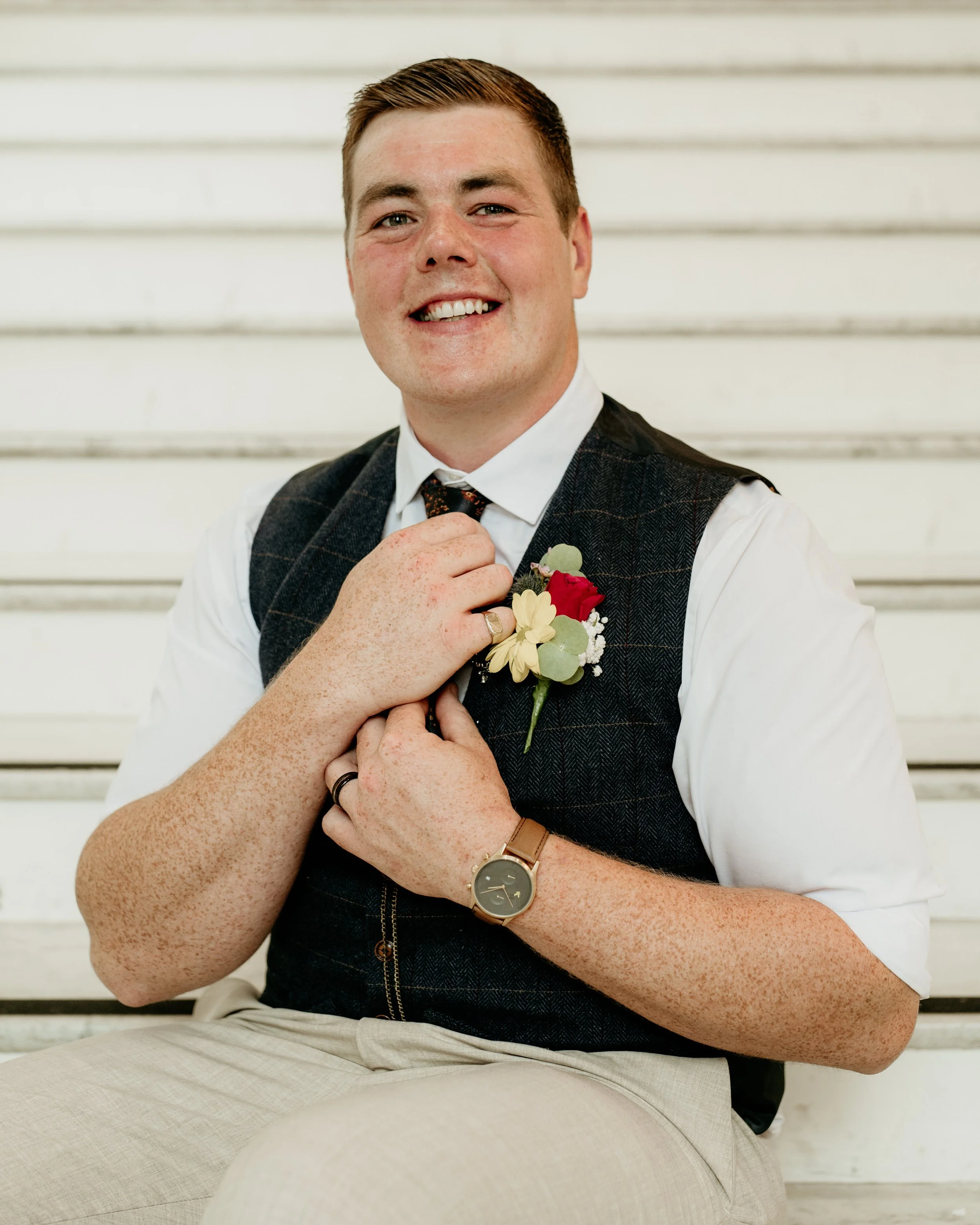 A groom sits on some steps and smiles at the camera while adjusting his tie.