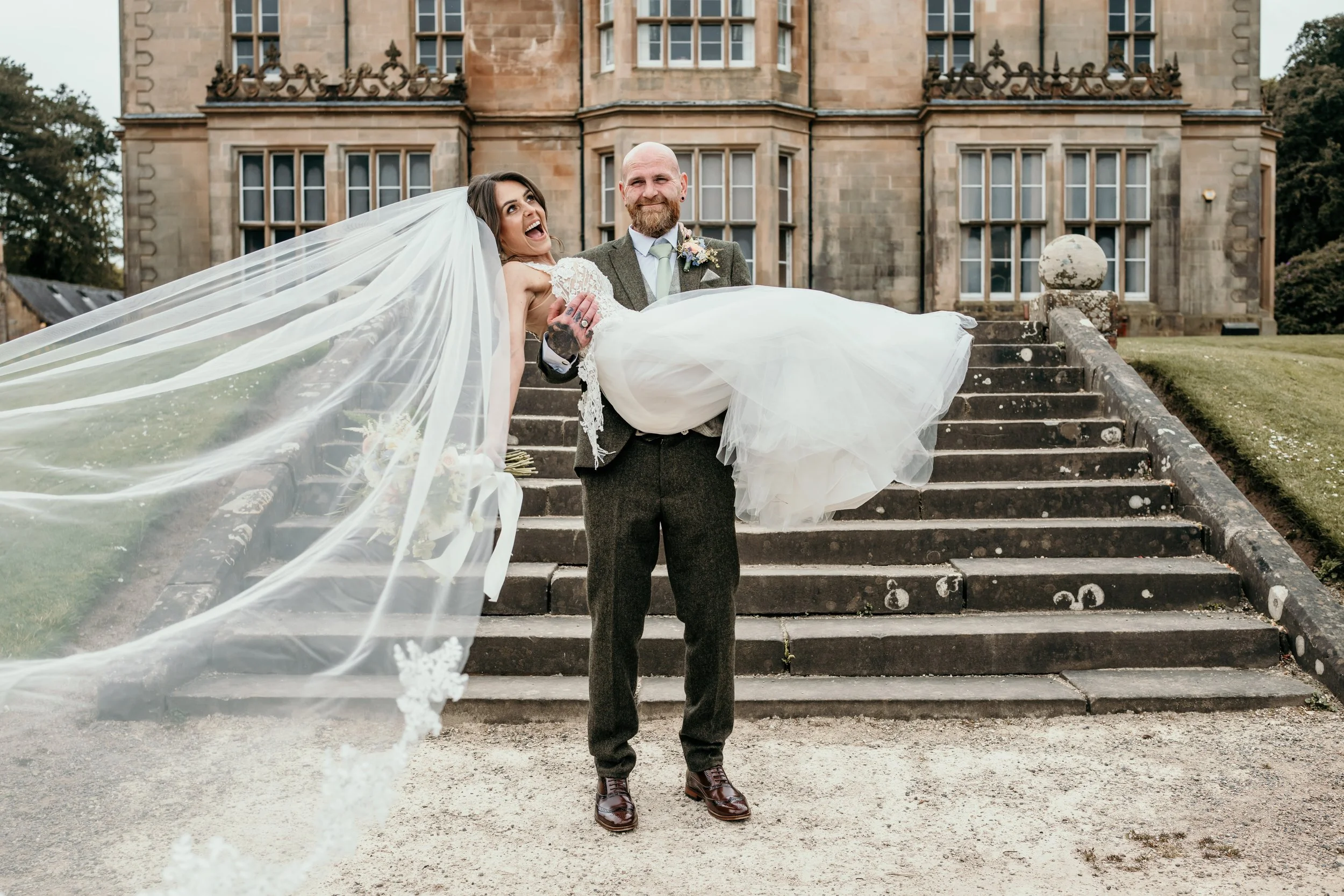 A newlywed couple standing on outdoor steps in front of a large historic stone building. The groom, with a beard, balding head, and wearing a gray suit with a tie, is carrying the bride. The bride, wearing a white lace wedding dress and a long veil, 