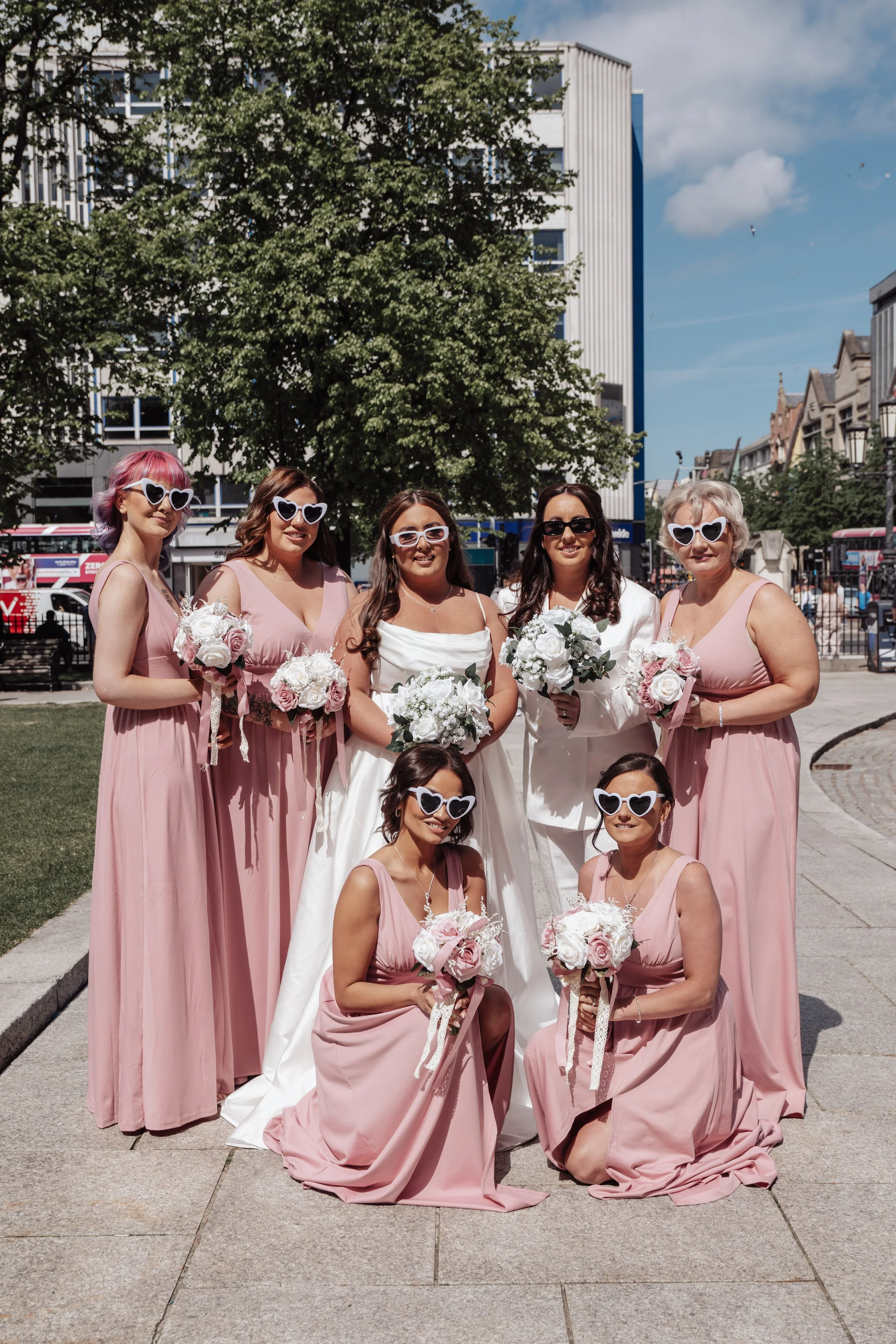 2 brides are surrounded by their bridesmaids on a sunny day outside Belfast City Hall. The are all wearing heart sunglasses and smiling at the camera while holding bouquets.