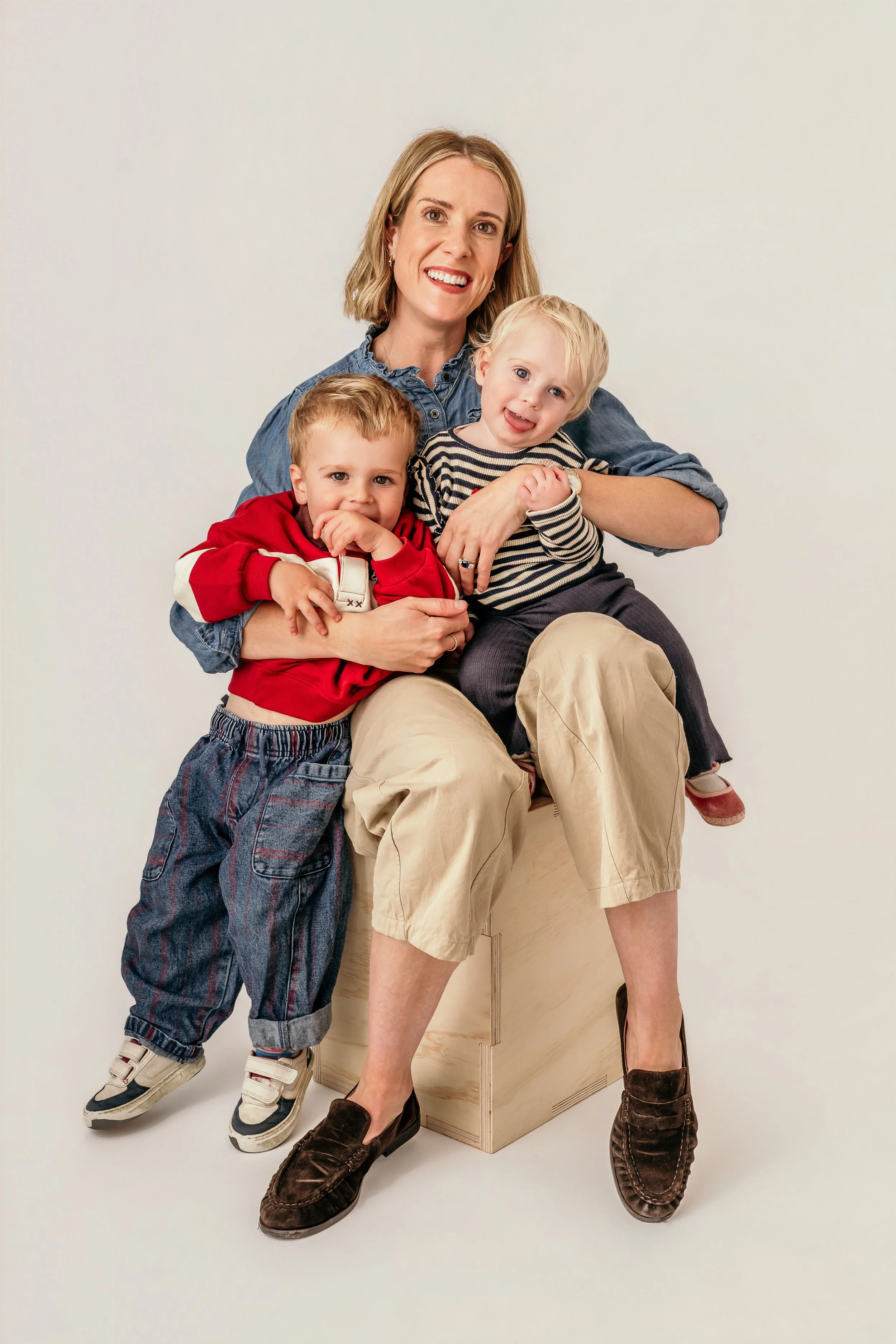 Mum in a photography studio sitting on a wooden block. Mum is holding two kids, a young boy and a young girl. All three are smiling at the camera.